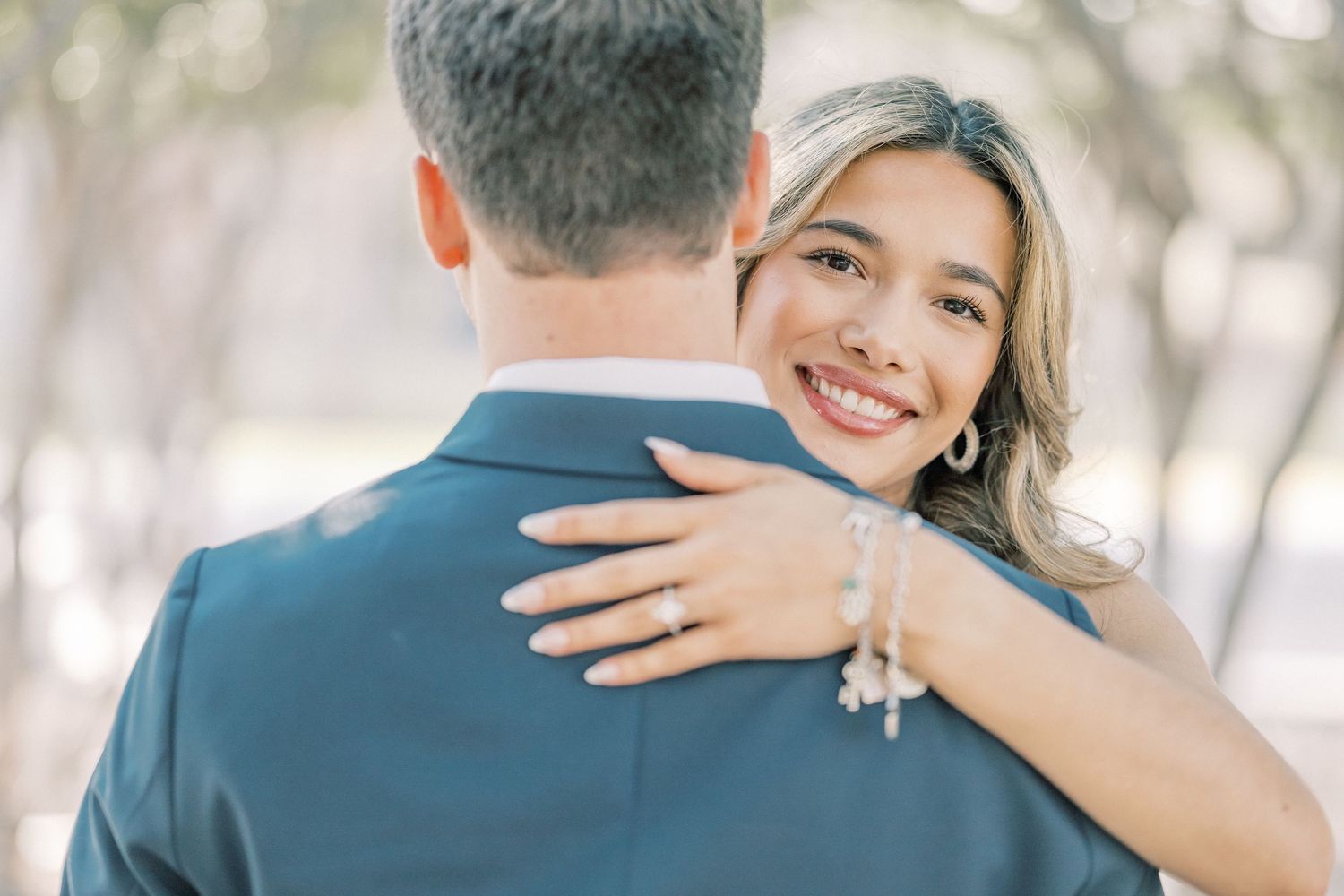 A couple embraces outdoors in a romantic moment with soft natural lighting filtering through trees in the background.