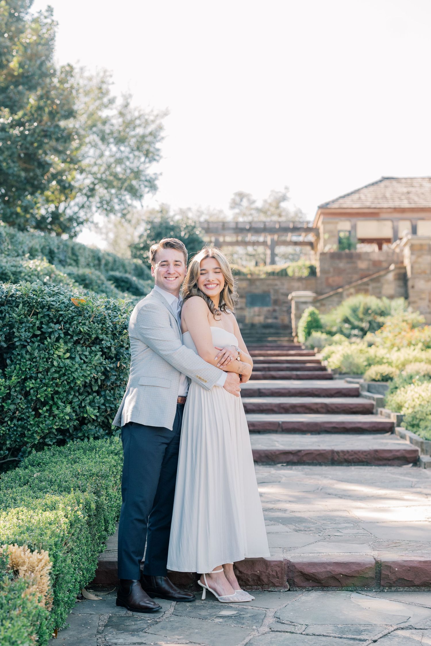 A romantic couple poses together on stone steps in a garden setting during golden hour.