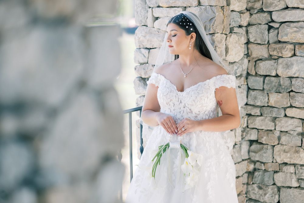 Dreamy side profile of bride in white lace gown holding flowers against stone architecture.