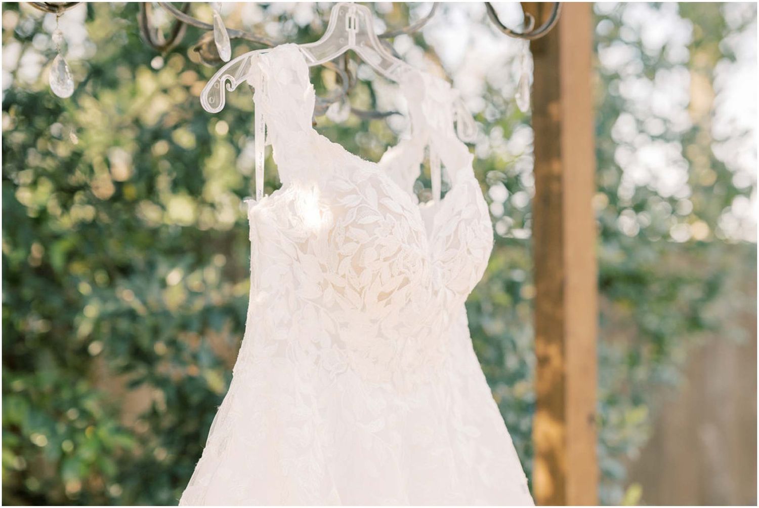A delicate white lace wedding dress hangs on a decorative hanger against a soft green outdoor background.