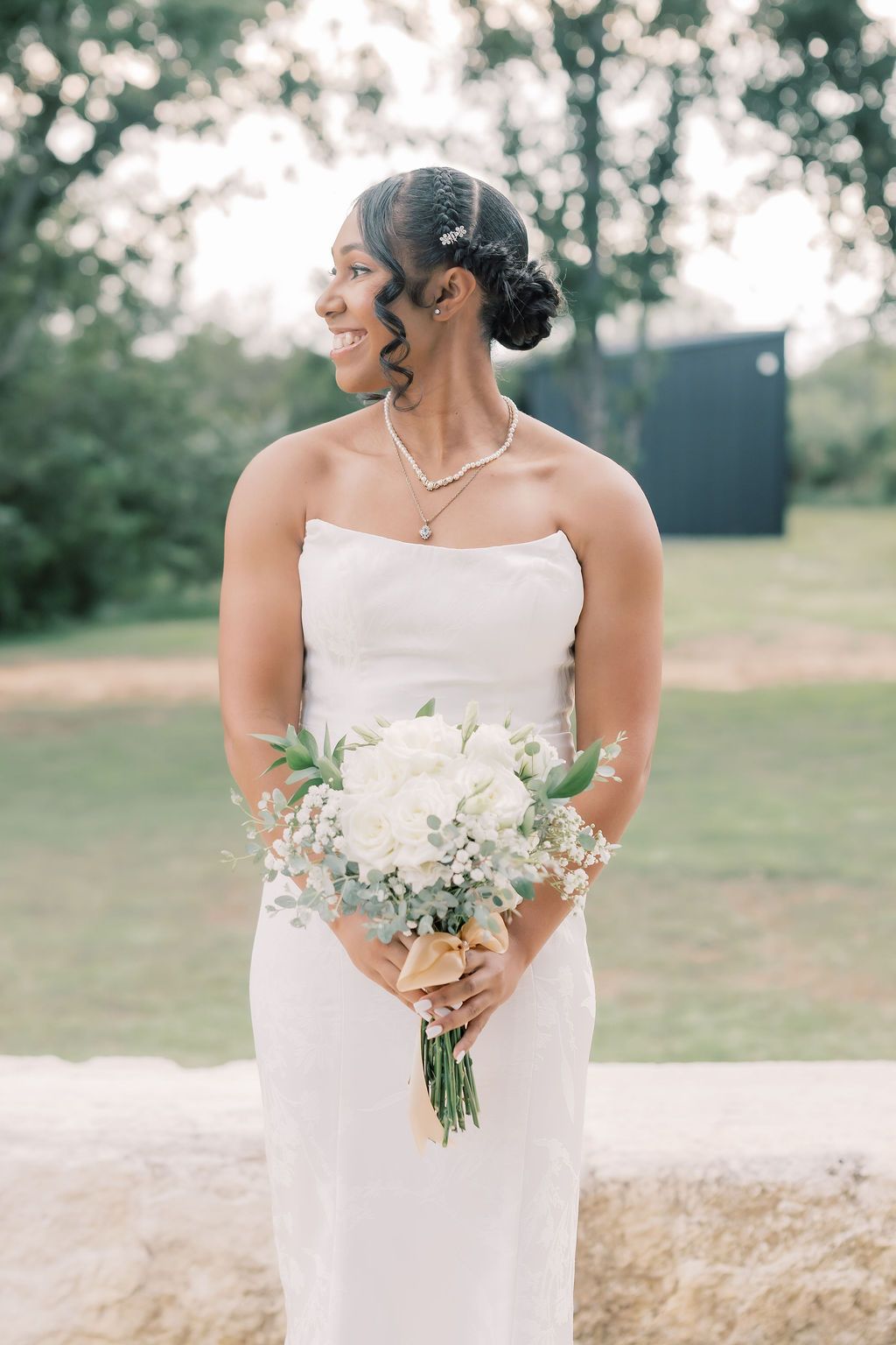 Smiling bride in strapless white gown holding white floral bouquet outdoors on her wedding day.