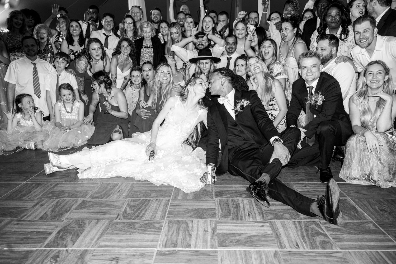 Black and white photo of bride and groom sharing a romantic dip kiss on the dance floor surrounded by cheering wedding guests.