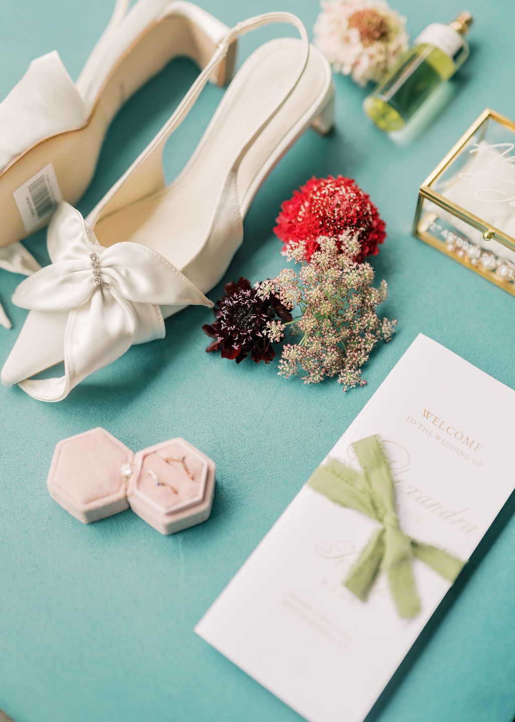 Elegant bridal flat lay with ivory bow heels, velvet ring box, floral bouquet, perfume, and green ribbon wedding program on teal background.