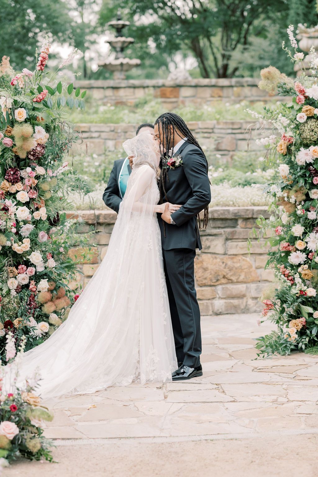 Bride and groom share first kiss at floral arch outdoor wedding ceremony on stone patio surrounded by lush pink blooms.