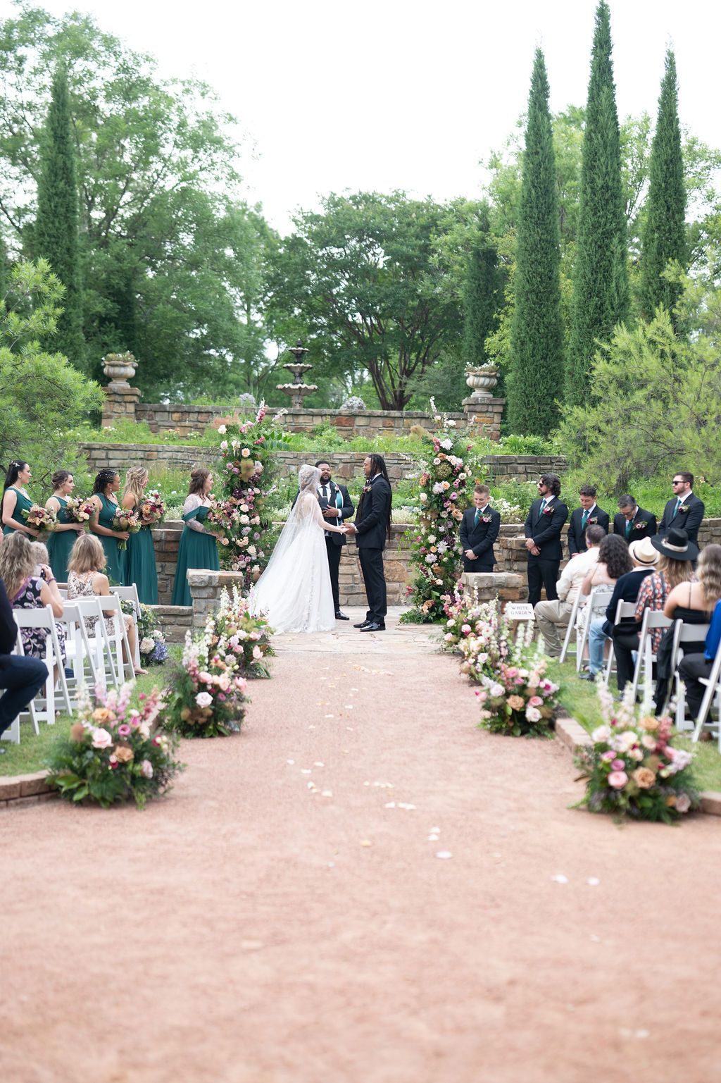 Outdoor wedding ceremony with floral aisle, arch, bridesmaids in green, and guests seated on white chairs amid lush greenery.