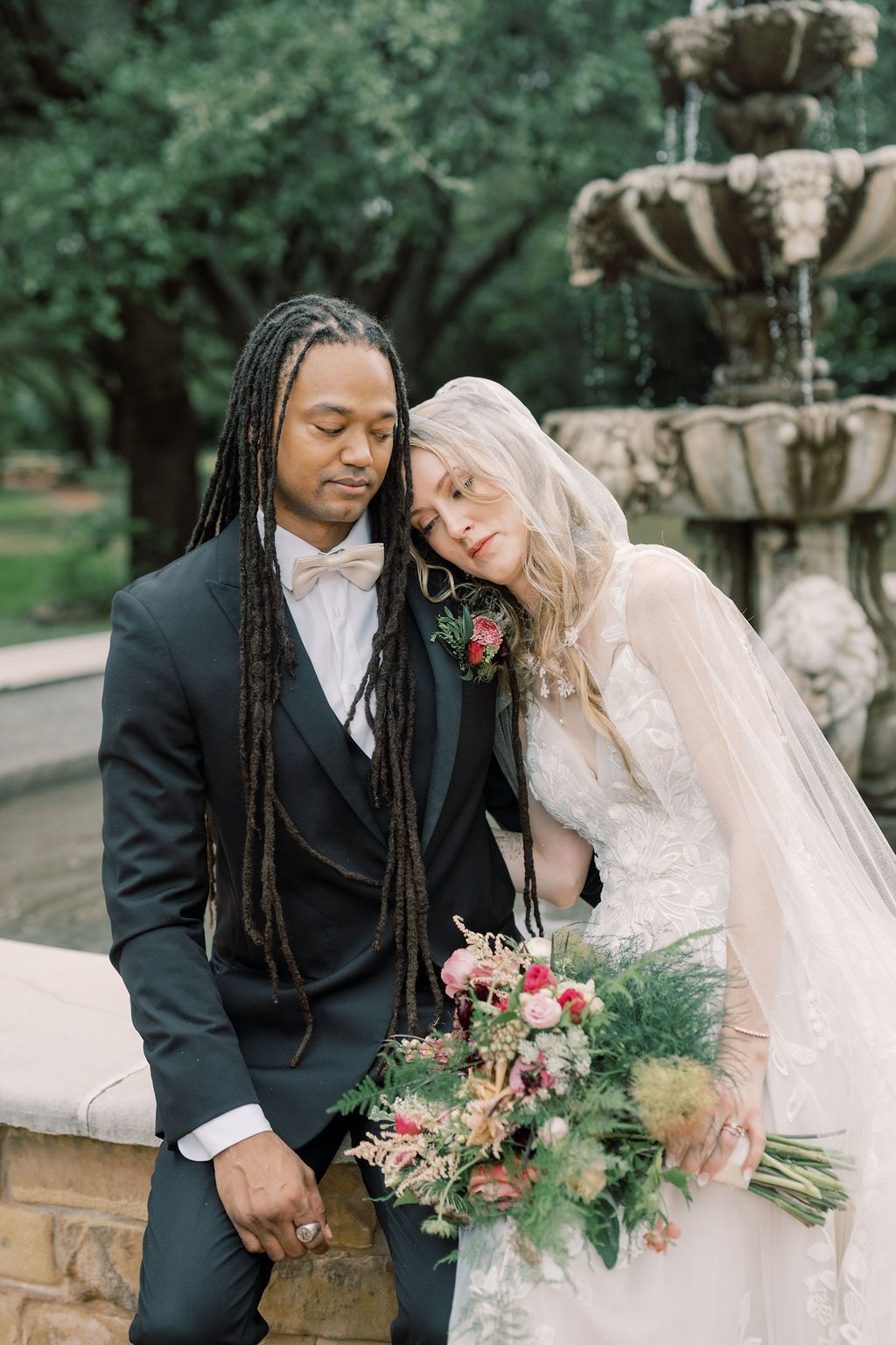 Interracial couple in wedding attire poses by stone fountain, bride in lace gown with bouquet leans on groom in black tuxedo.