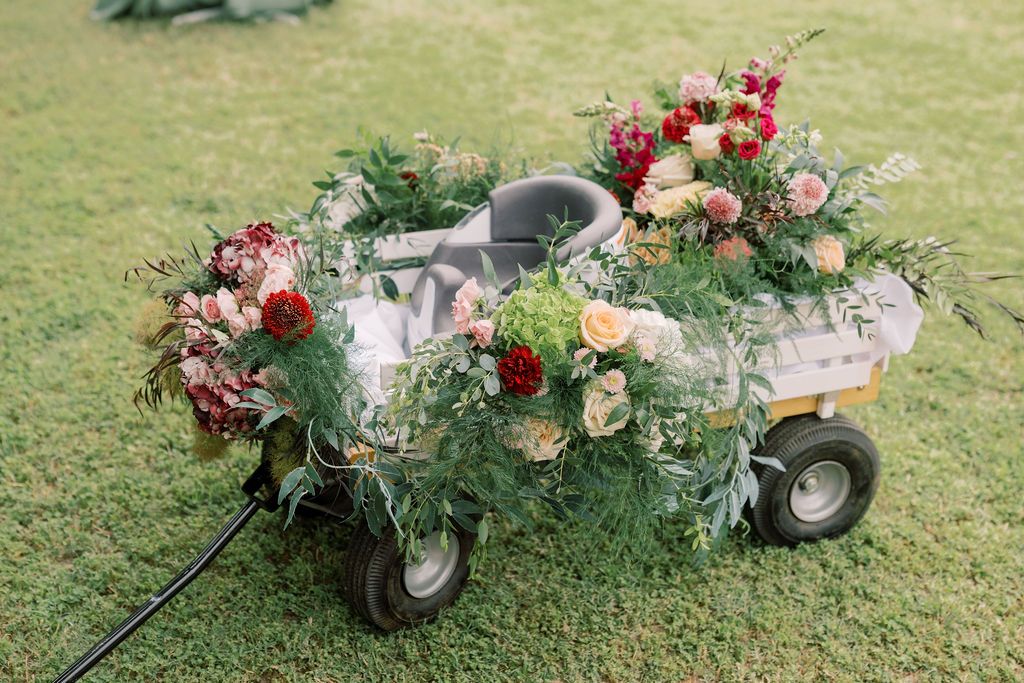 A decorative wagon overflowing with lush greenery and colorful blooms including red, pink, and cream flowers on green grass.