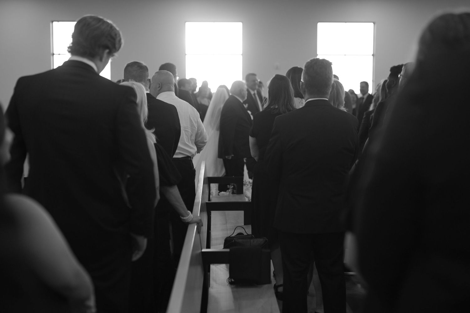 Wedding guests standing in church pews during ceremony with bright light from windows.