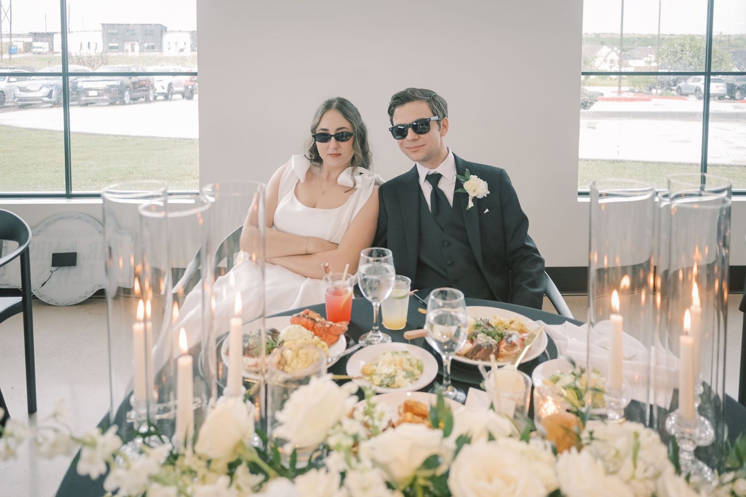 Bride and groom wearing sunglasses seated at sweetheart table with candles and floral centerpiece.