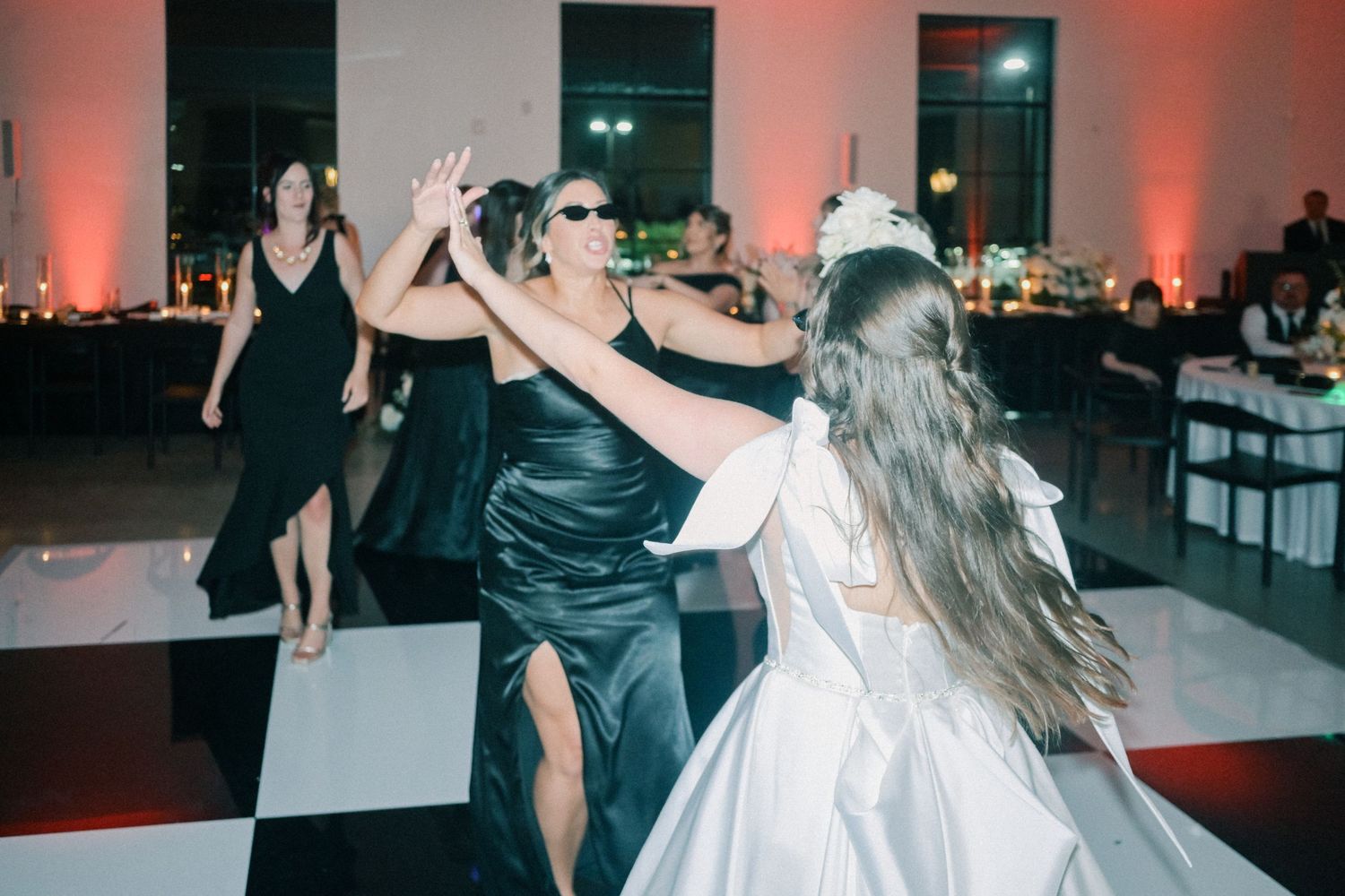 Bride dances joyfully with guests in black dresses on a checkered dance floor.