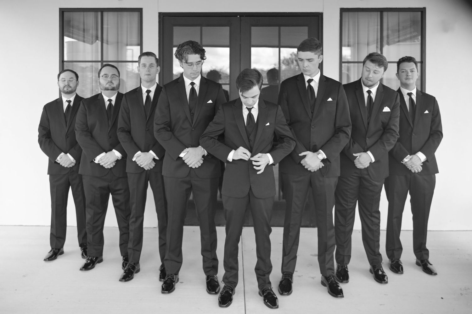 Groom and groomsmen posing in black suits outside a white building in a black and white photo.