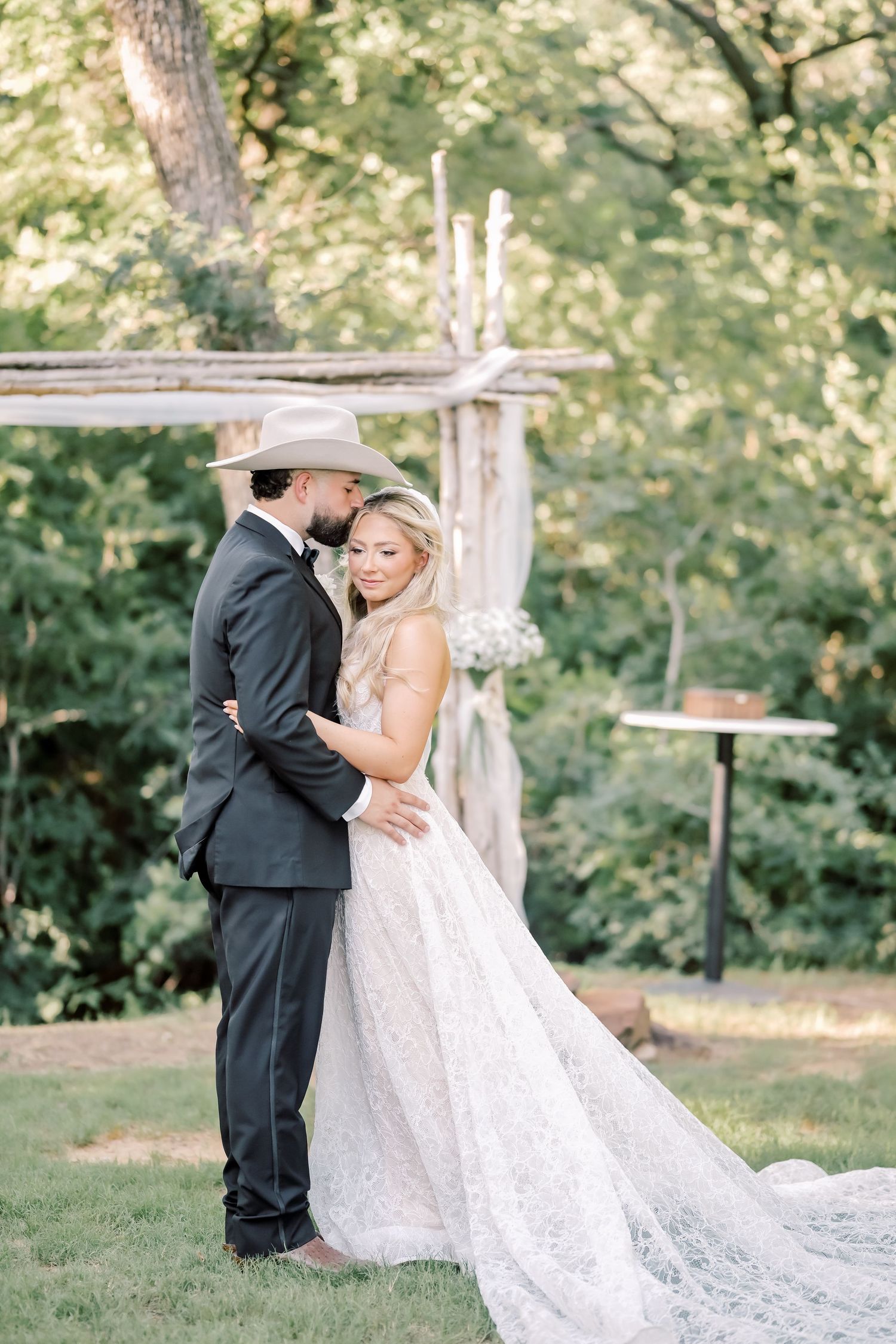 Bride and groom embrace outdoors under rustic wooden arch surrounded by lush greenery at elegant wedding ceremony.