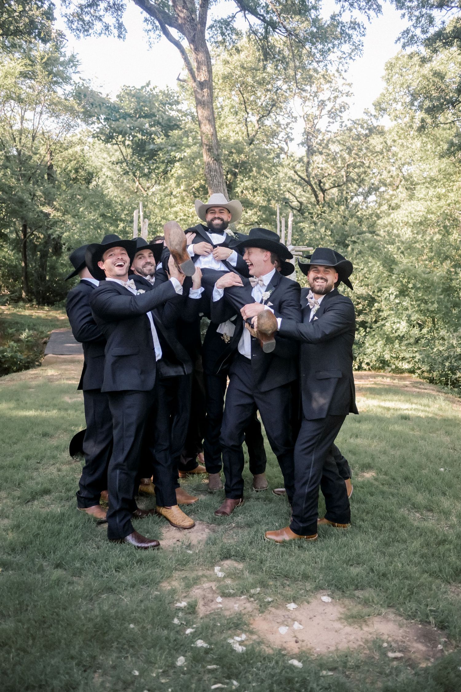 Groomsmen in black suits and cowboy hats playfully lift groom in outdoor wooded wedding celebration.