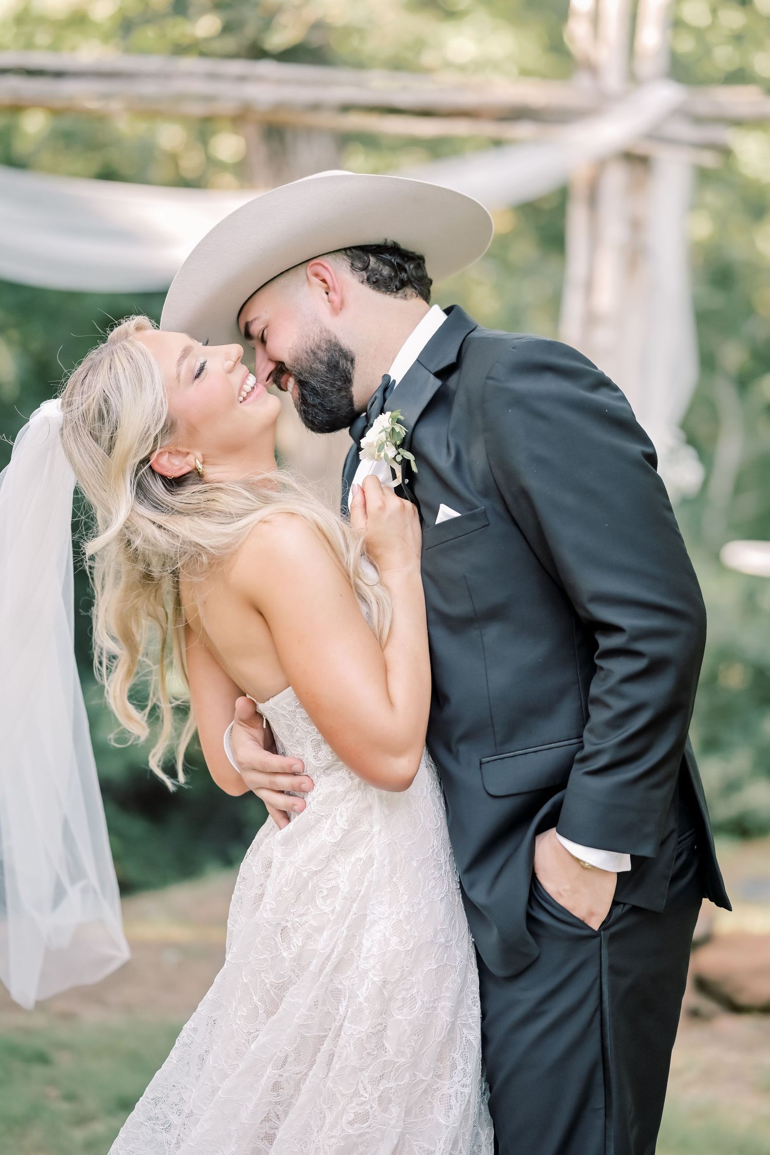 Bride and bearded groom in white cowboy hat share a joyful kiss at outdoor wedding ceremony.