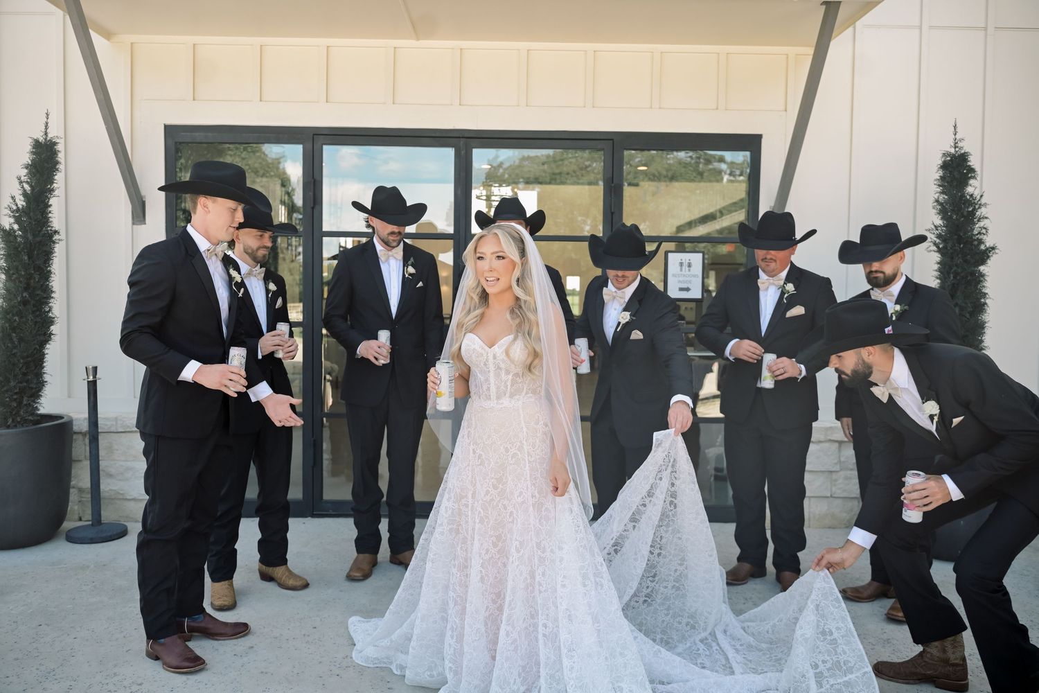 Bride in lace gown surrounded by groomsmen in black cowboy hats holding her train outside a venue.