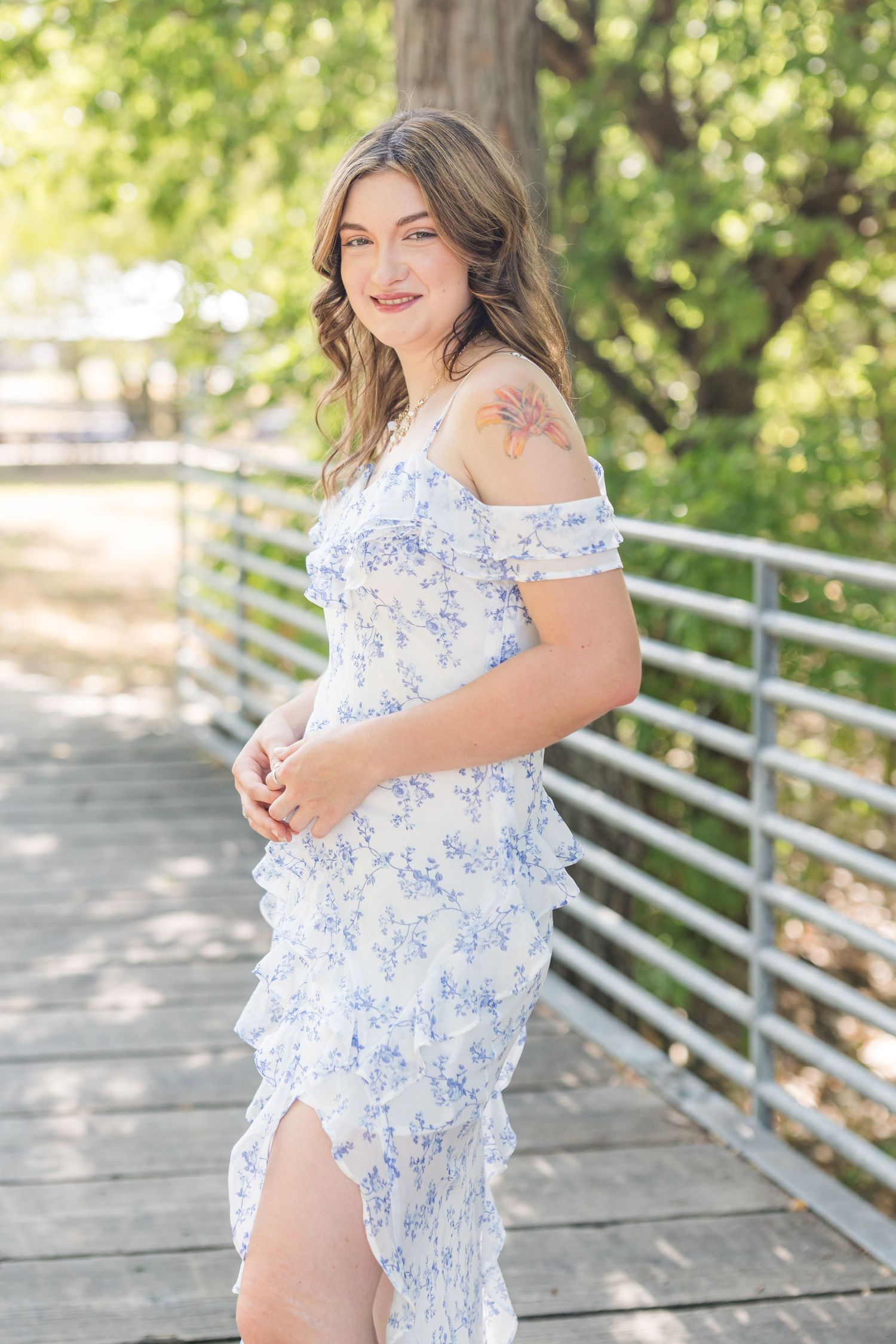 Young woman in white floral off-shoulder dress poses on a bridge surrounded by lush green trees.