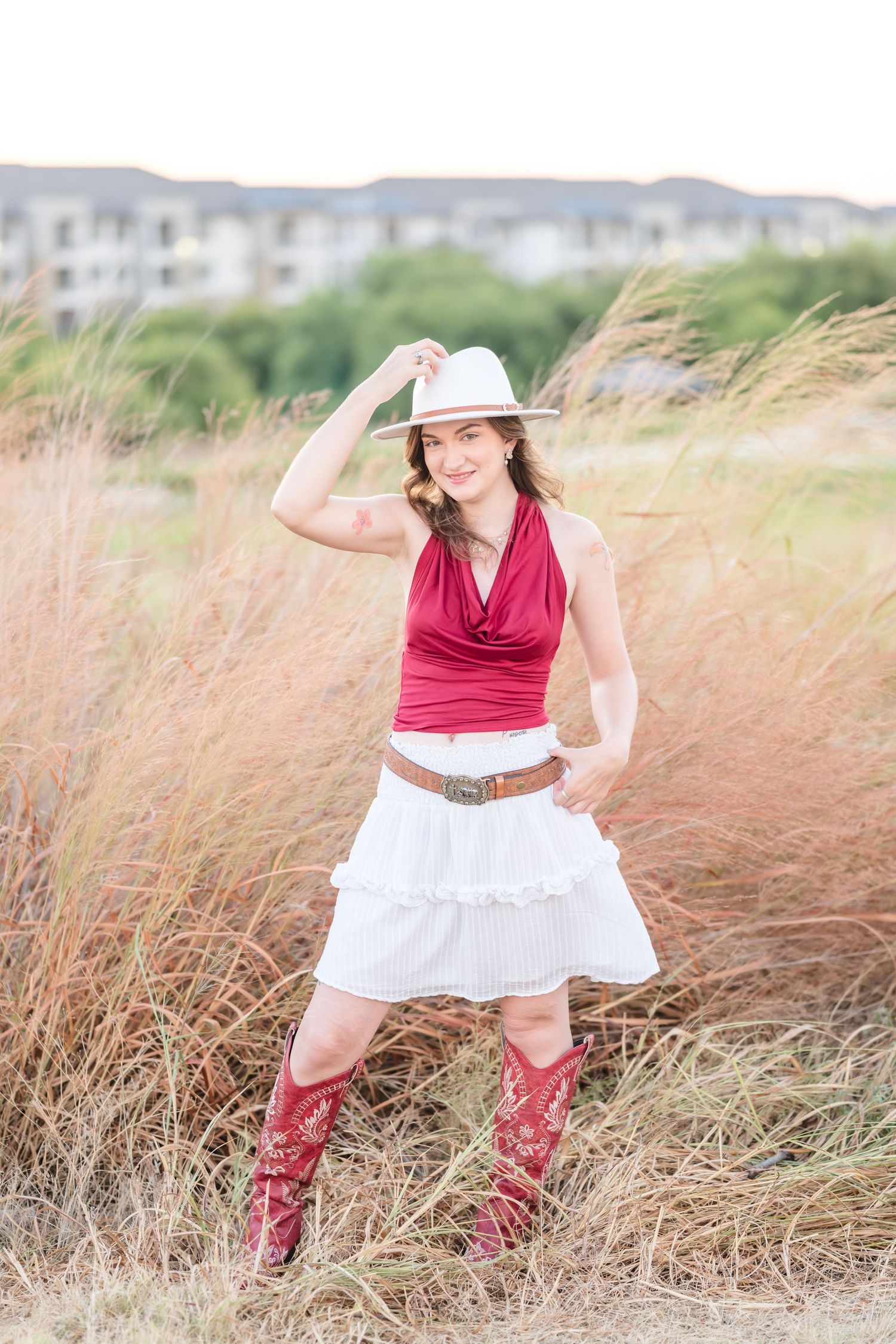 Young woman in red halter top, white ruffled skirt, red cowboy boots, and white hat posing in tall grass field.