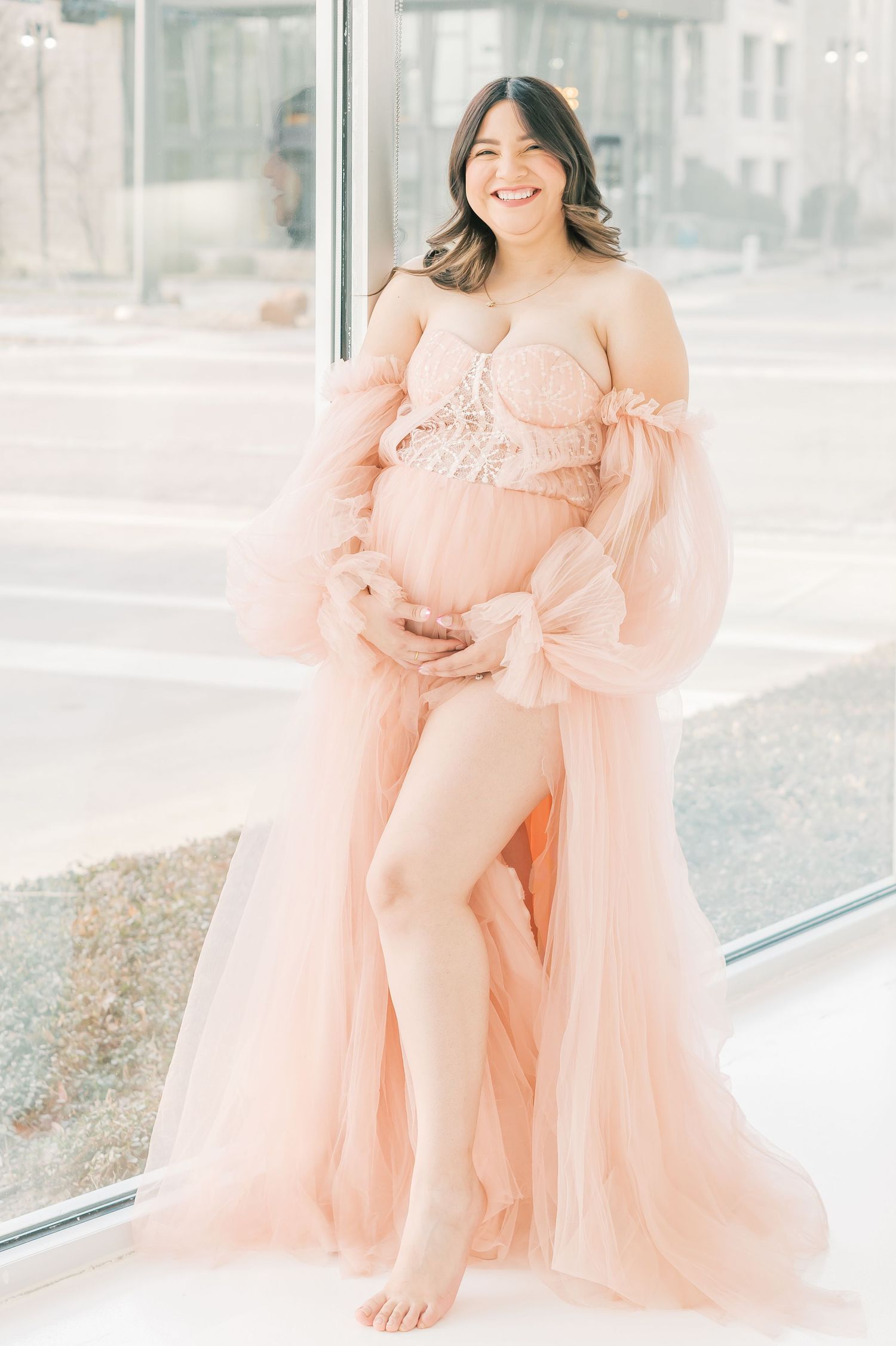 Smiling pregnant woman in pink tulle maternity gown holding baby bump near large window.