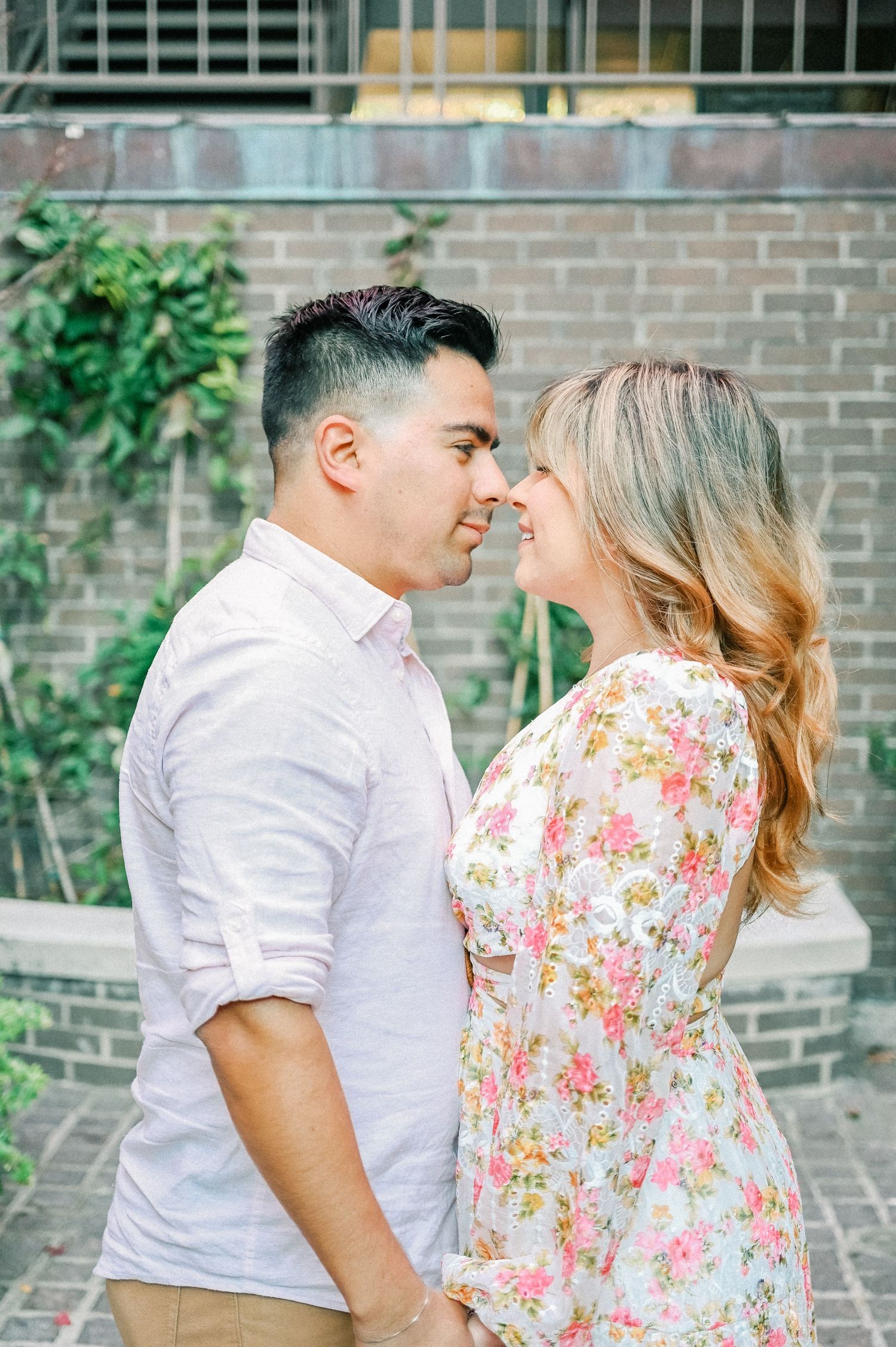 Couple in light spring outfits gazing into each others eyes in a lush brick courtyard.