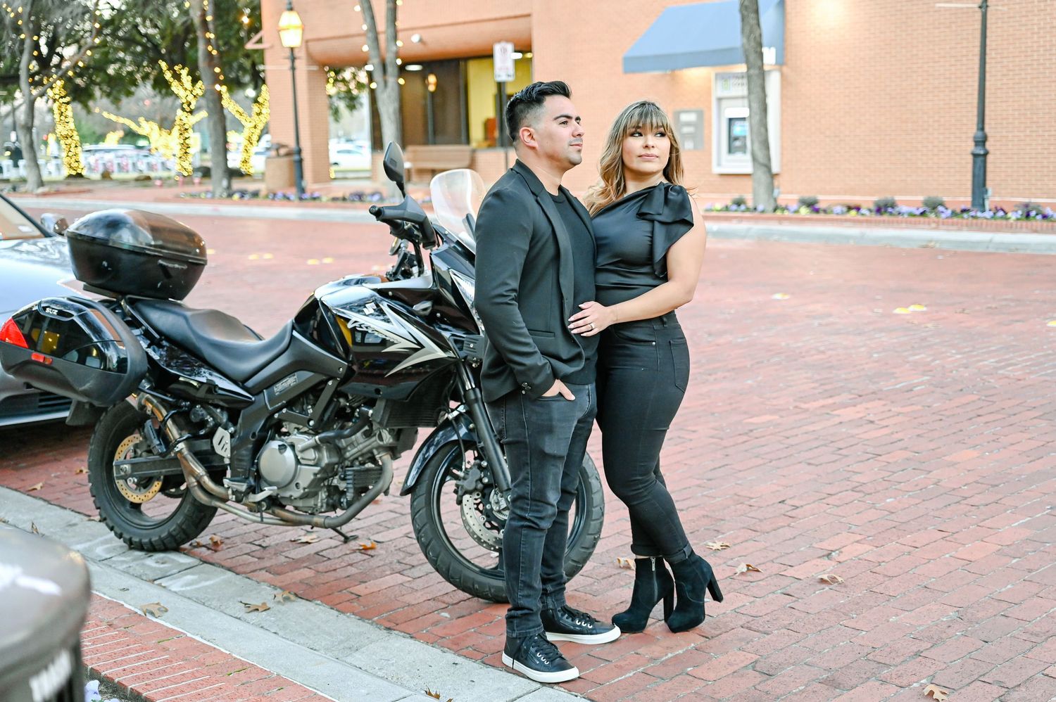 A stylish couple dressed in all-black outfits posing beside a black motorcycle on a brick sidewalk in an urban setting.