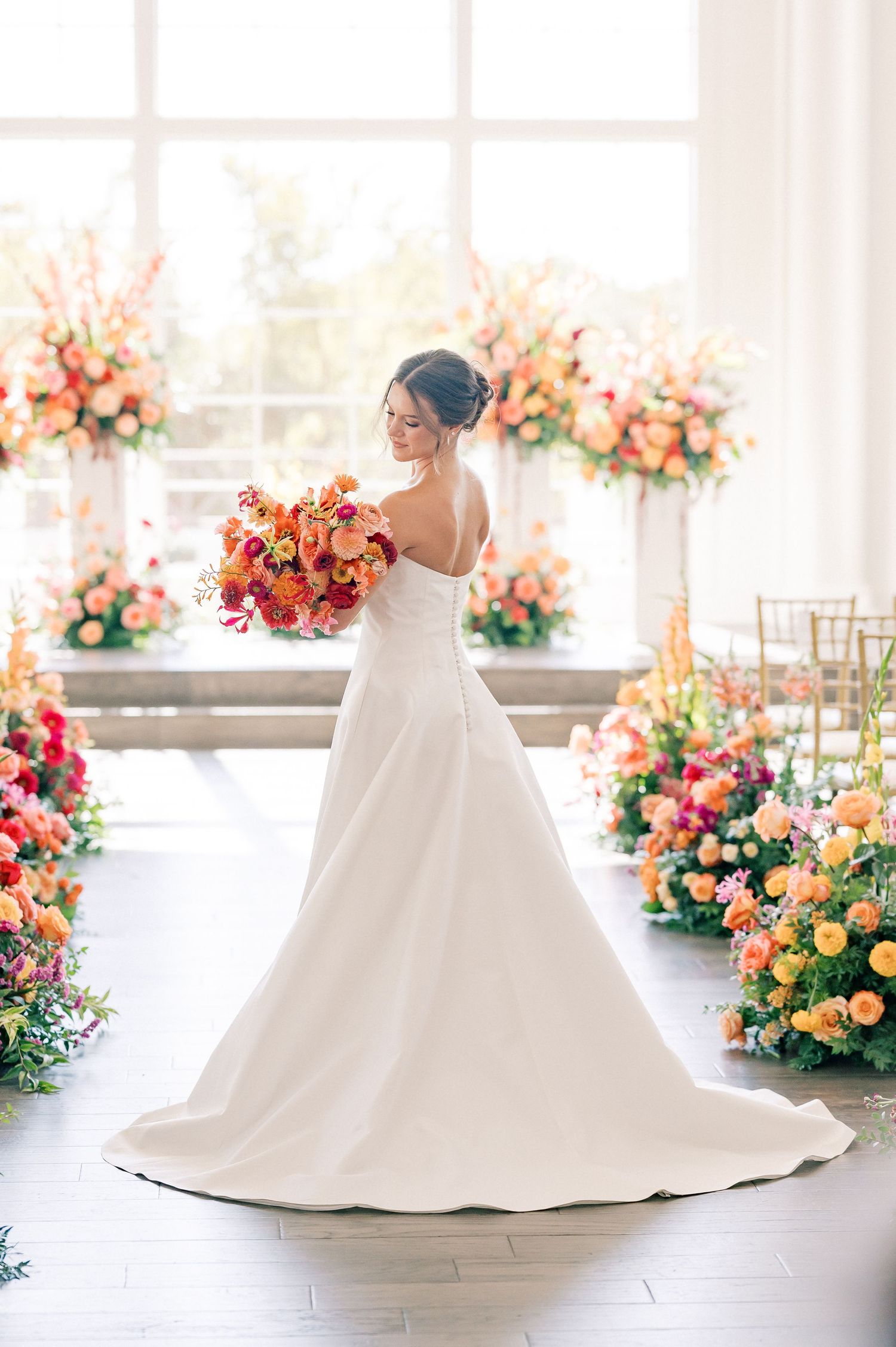 Bride in white strapless gown holding vibrant orange and pink bouquet, surrounded by lush floral arrangements in bright venue.