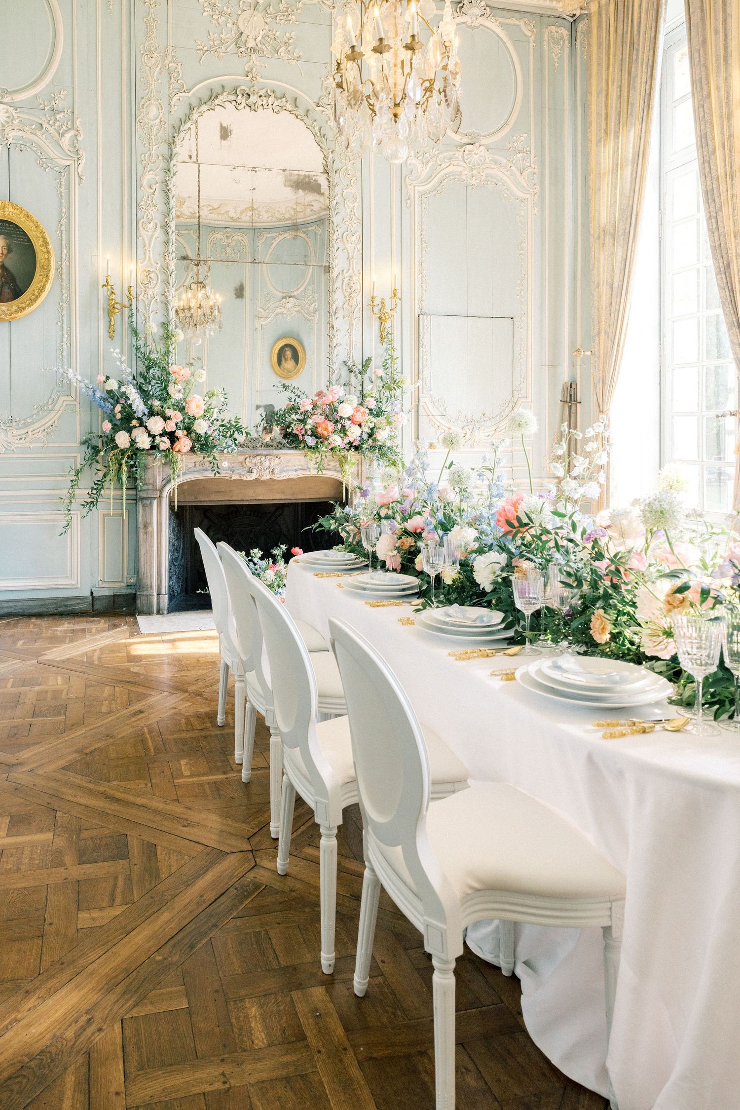 Elegant French wedding reception with white chairs, floral garland table runner, ornate mirror, and chandelier in a pastel ballroom.