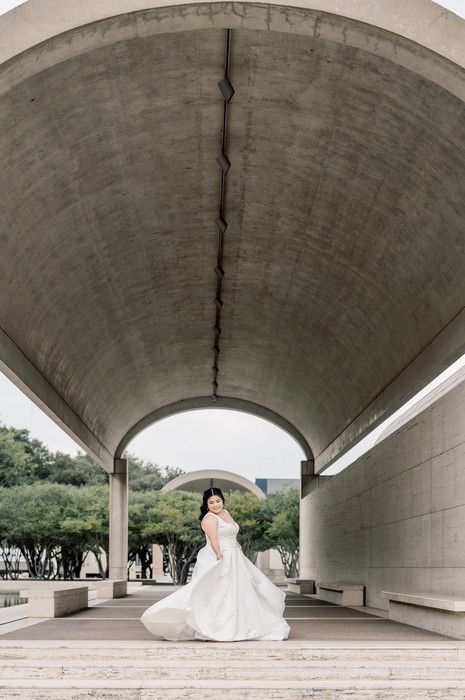 Bride in white gown twirling under curved concrete archway at modern architectural venue.