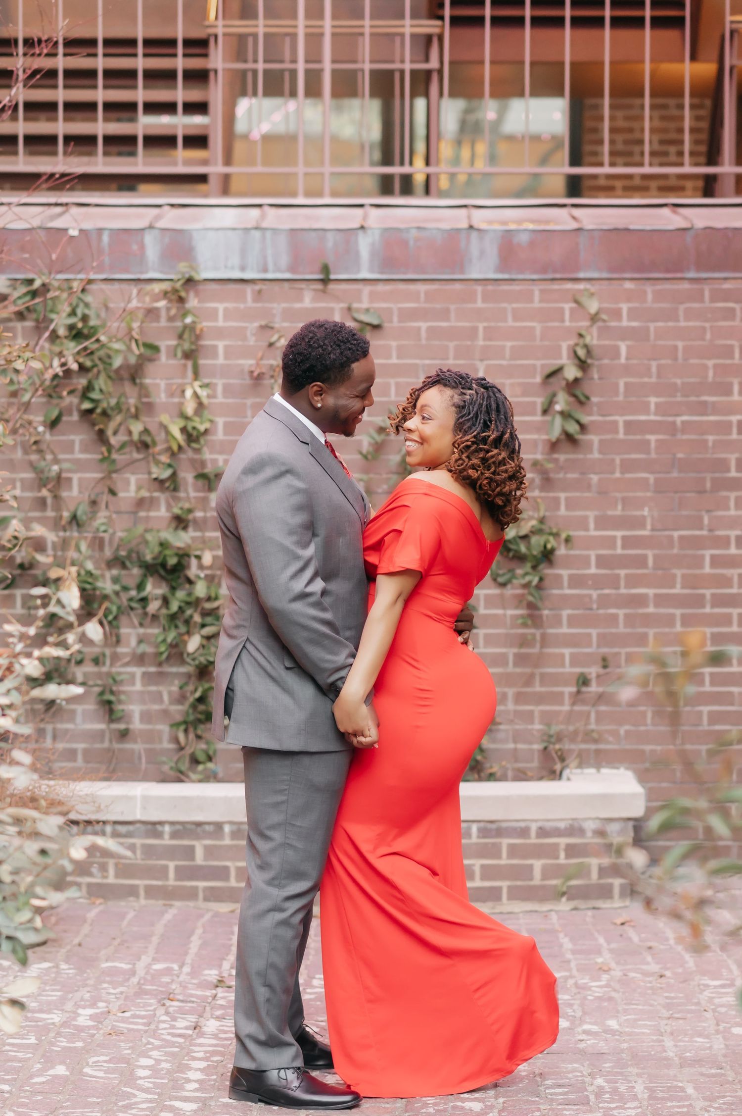 Couple in elegant red gown and gray suit embracing and smiling in front of a brick wall with ivy.