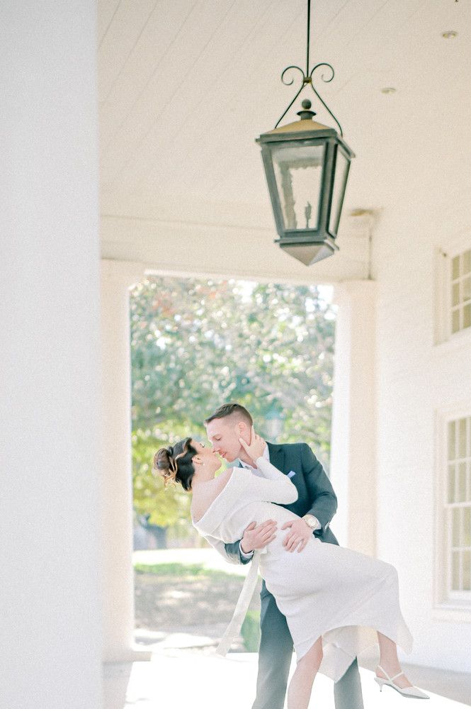 Groom dips bride for a romantic kiss under a lantern on a white columned porch on their wedding day.