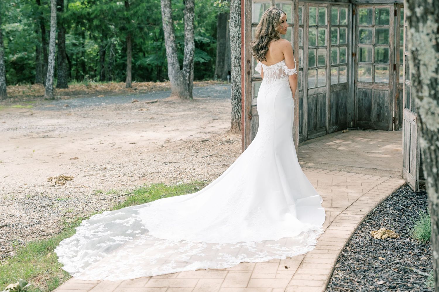 Bride in off-shoulder white mermaid wedding gown with lace train standing at rustic outdoor venue.