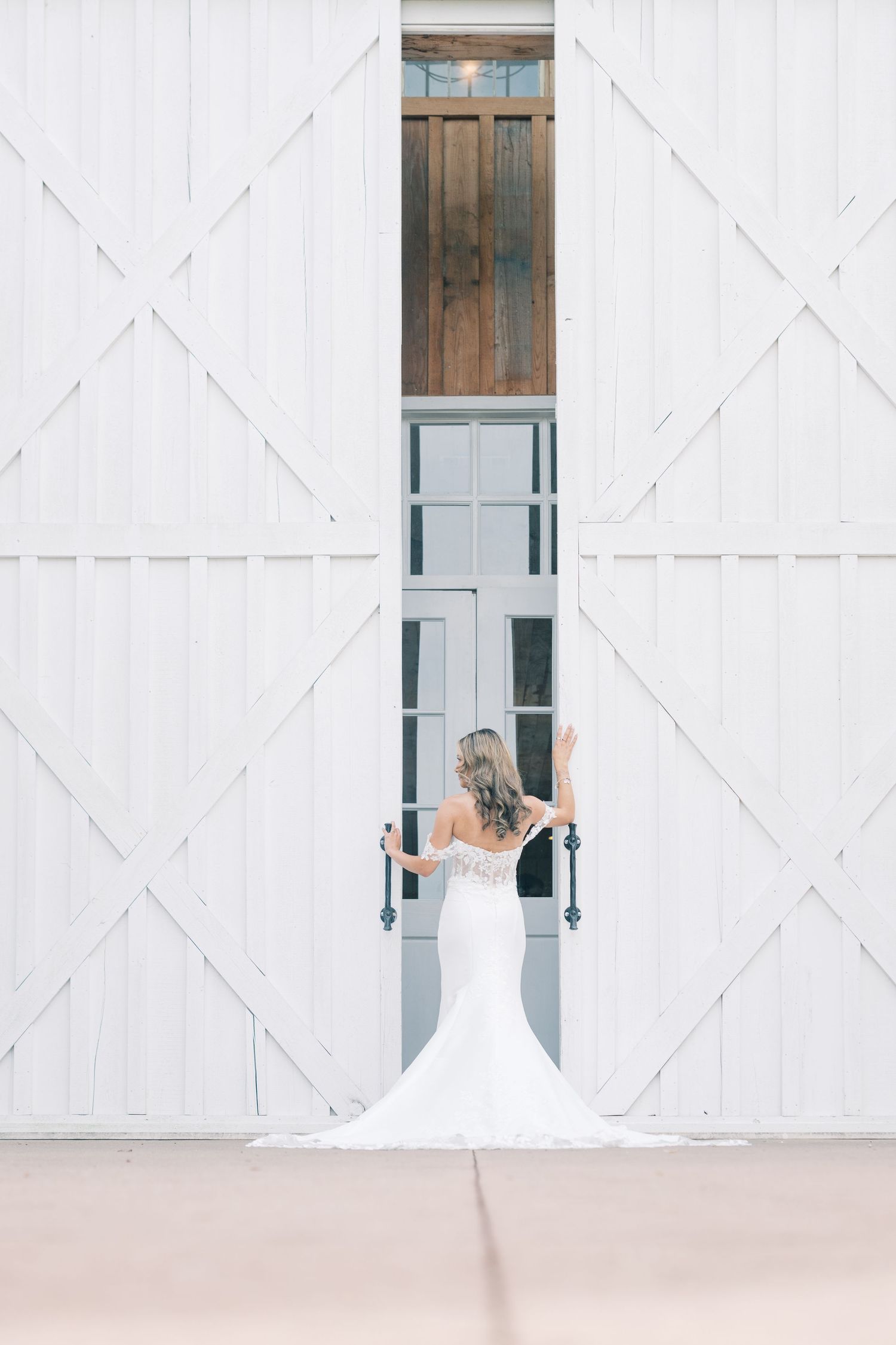 Bride in mermaid wedding gown standing before large white barn doors with arms raised at wedding venue.