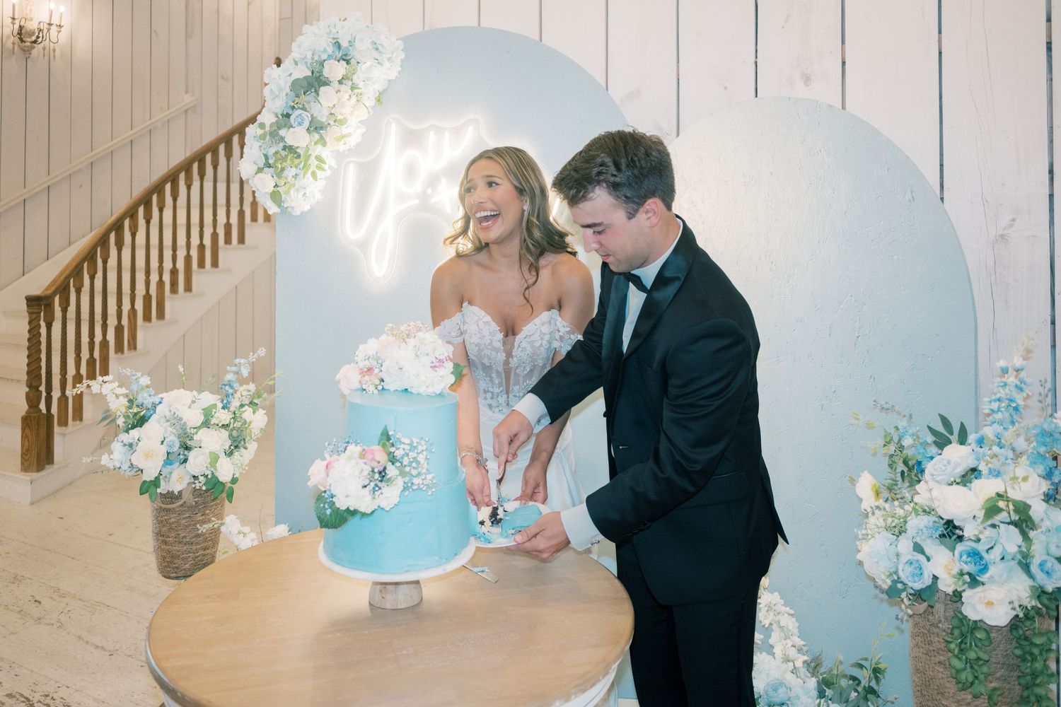 Bride and groom cutting light blue wedding cake at reception with floral arch backdrop and staircase.