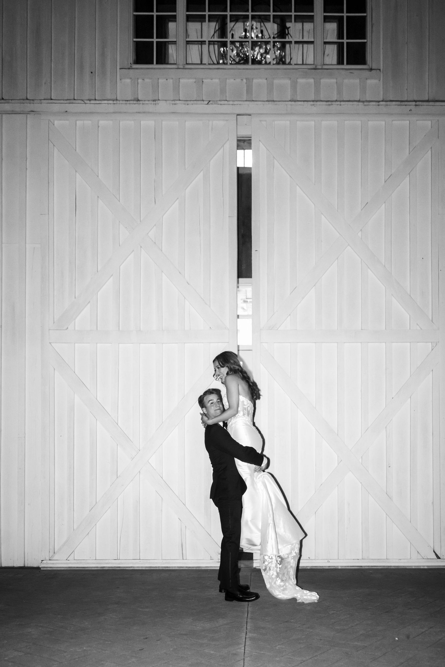 Black and white photo of groom lifting bride in front of large white barn doors at wedding venue.