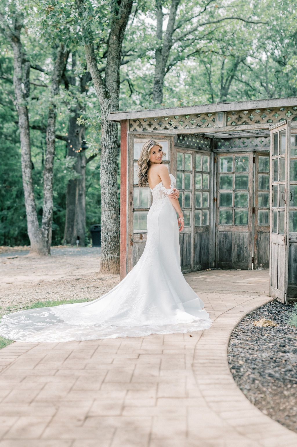 Bride in elegant white off-shoulder mermaid gown with long train poses at rustic wooden pergola surrounded by lush trees.