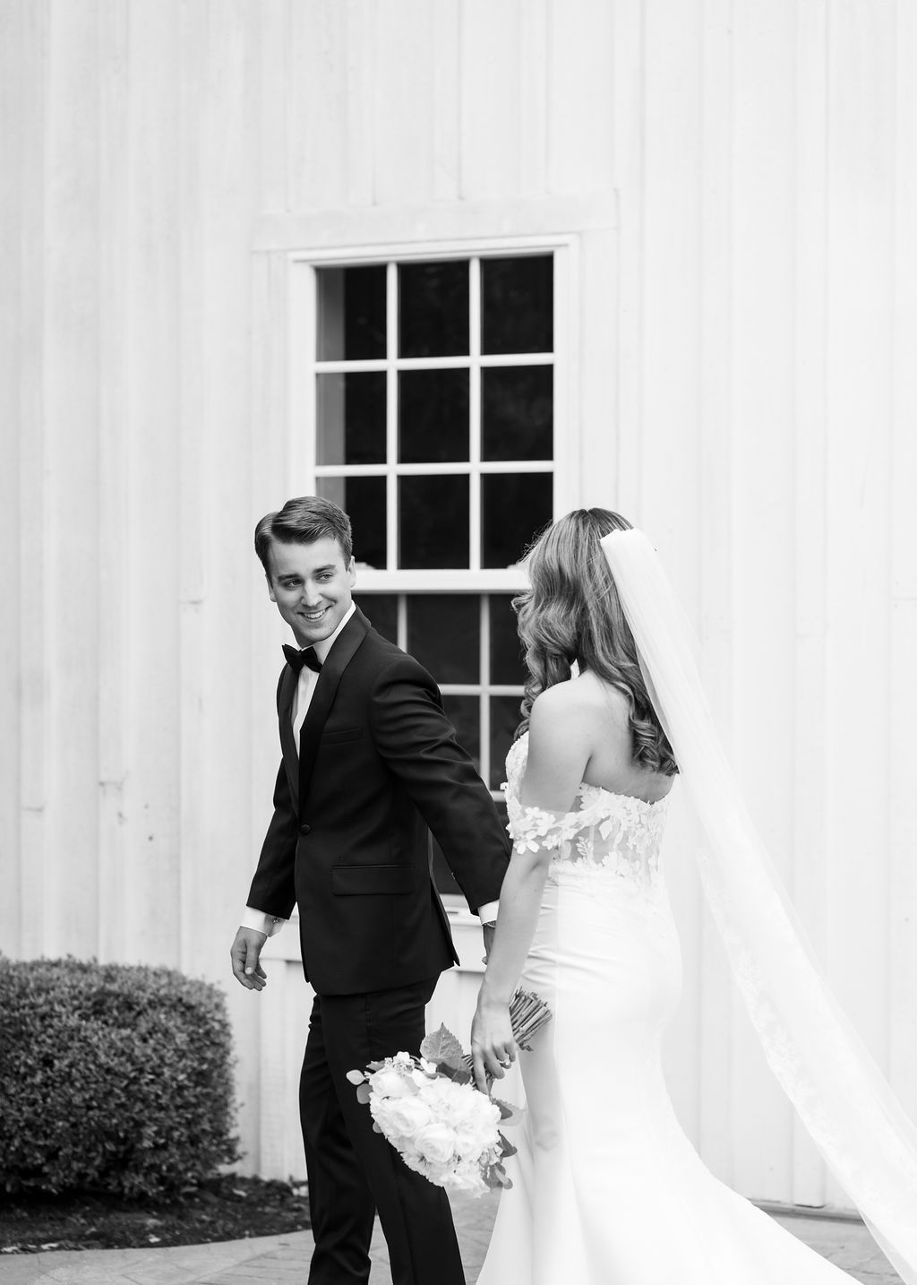 Groom smiles back at bride in elegant black tuxedo as she follows in a fitted wedding gown with veil.