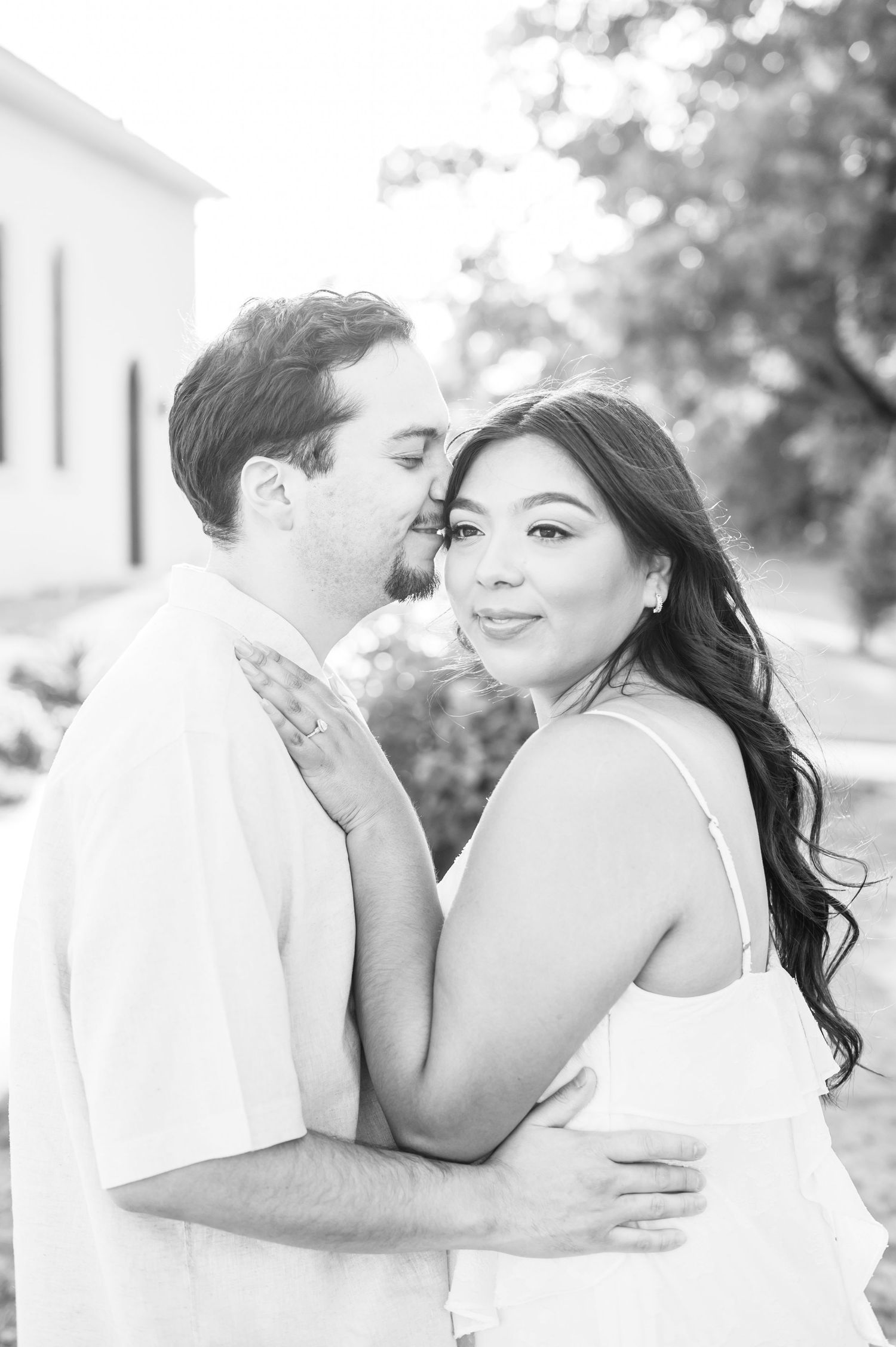 A romantic black and white portrait of a couple embracing outdoors with soft bokeh lighting in the background.