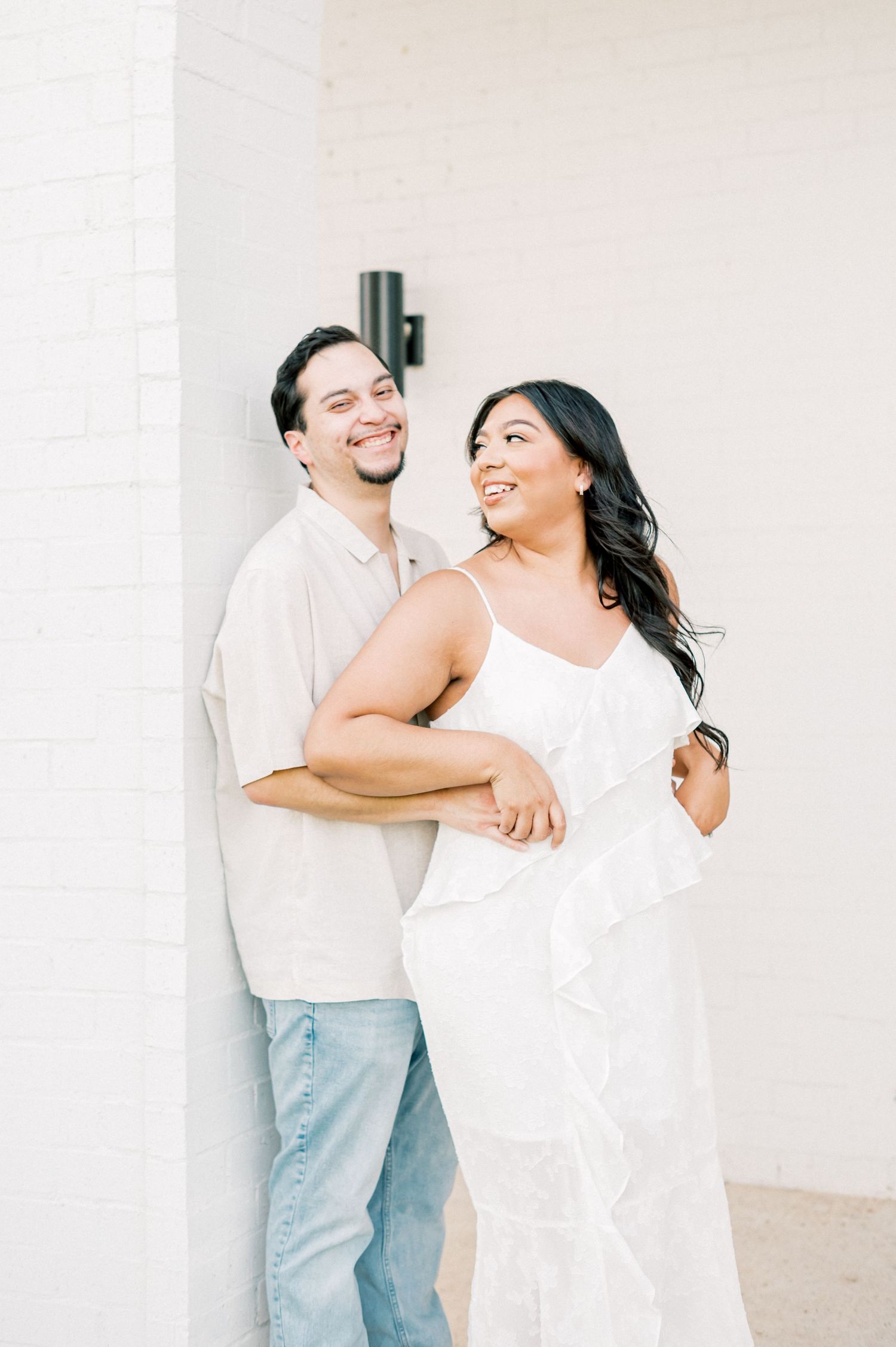 A couple dressed in white share joyful embraces against a light colored wall backdrop.