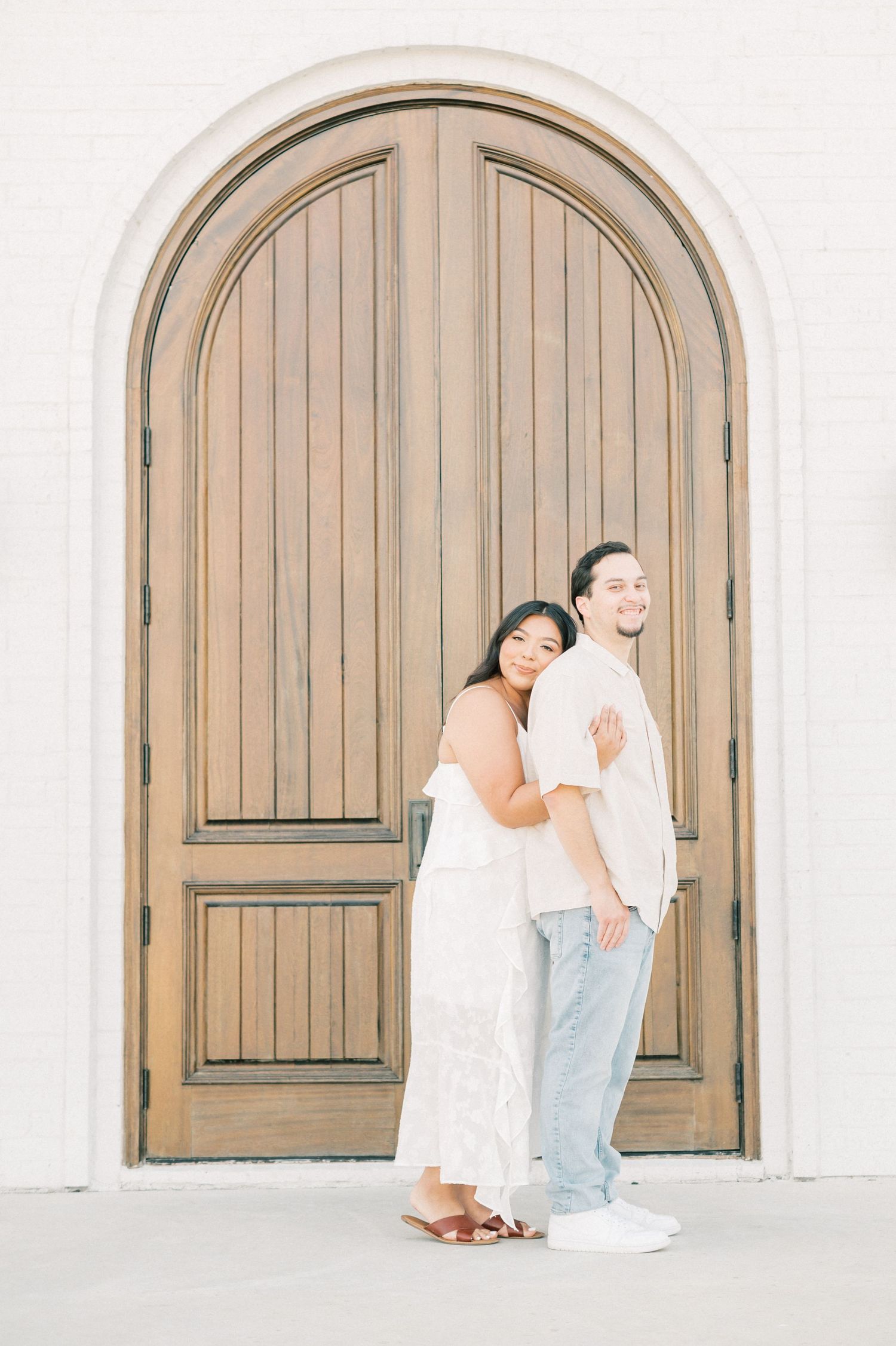 A couple shares a playful moment against a white stucco wall with an arched wooden door in the background.