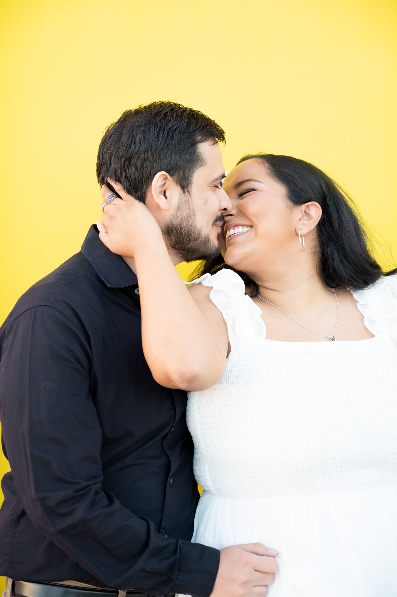 Couple in black and white outfits embracing lovingly against yellow wall.