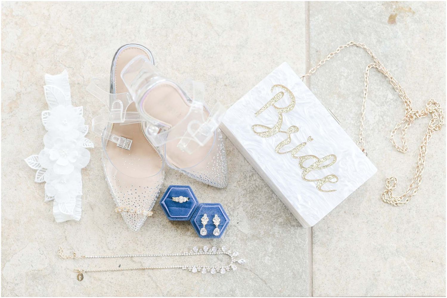 Bridal flat lay featuring clear heels, lace garter, blue hexagon earrings, gold necklace, and white Bride jewelry box.