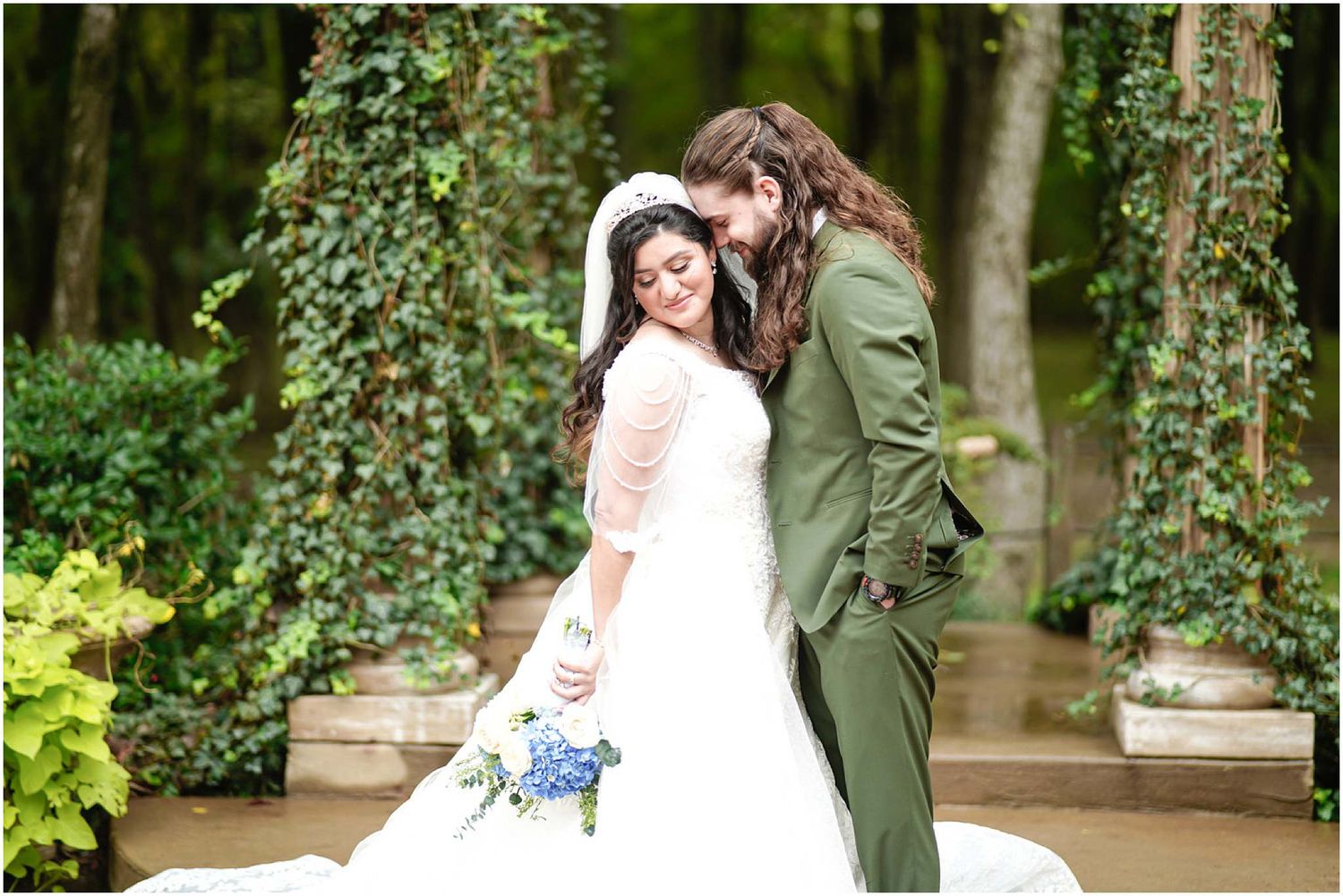 Bride and groom embrace in lush garden setting, groom in green suit nuzzling bride in white gown with blue bouquet.