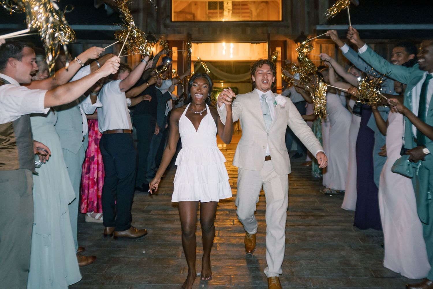 Newlyweds exit through a sparkler send-off tunnel, bride in white mini dress and groom in beige suit.
