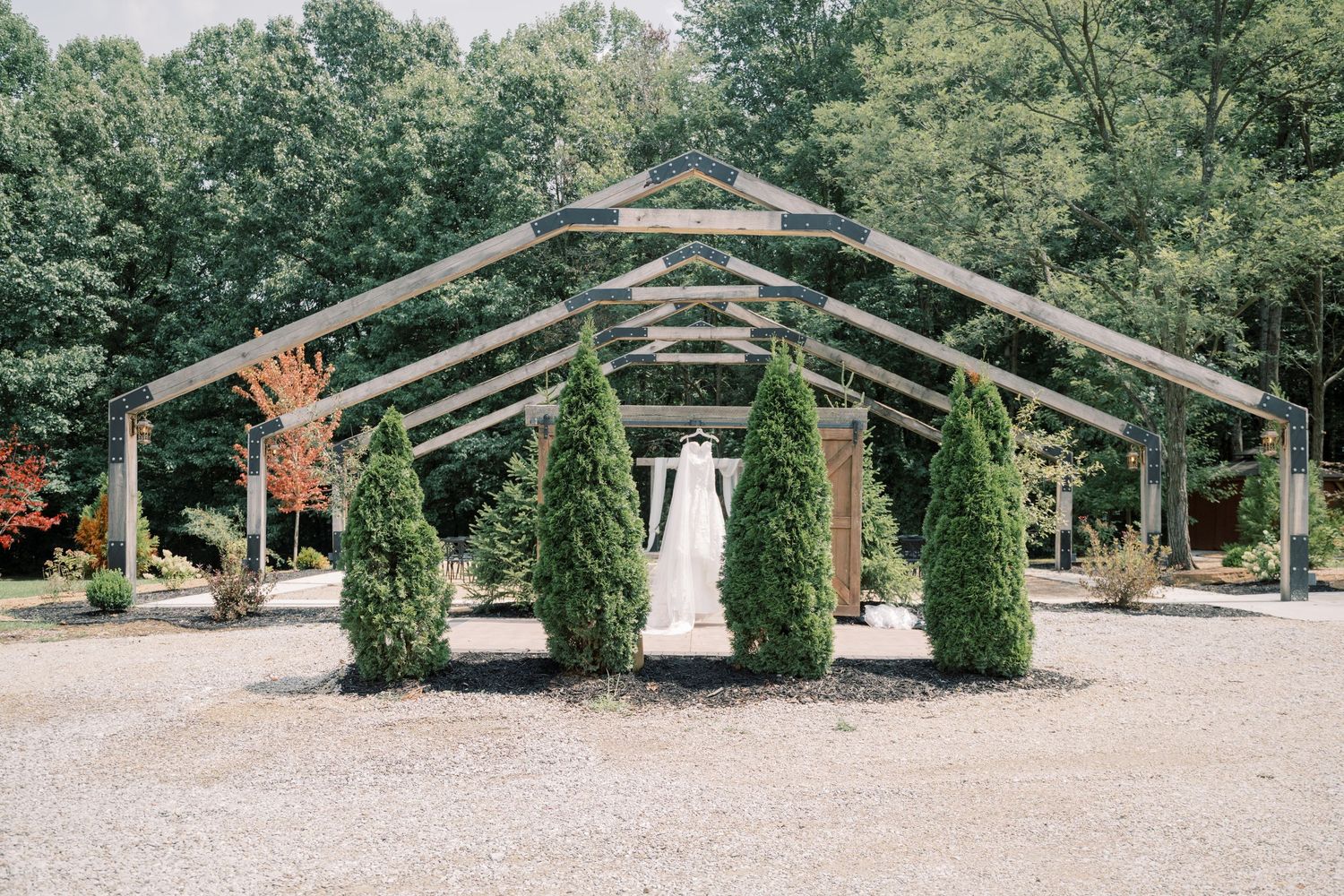 Bride stands under rustic wooden pergola surrounded by evergreen trees at an outdoor wedding venue.