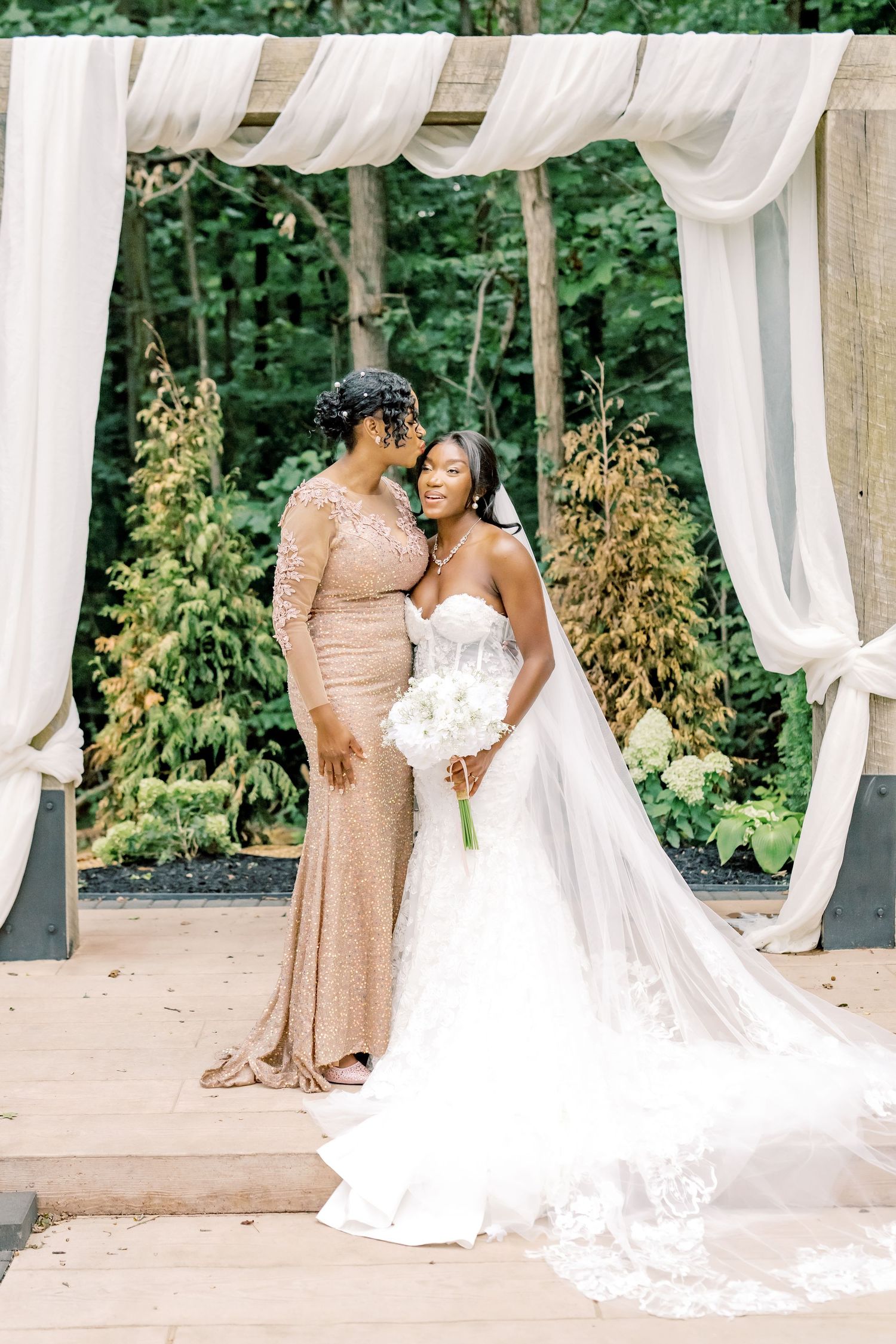 A bride in a white gown holds a bouquet beside a woman in a gold dress under a draped outdoor wedding arch.