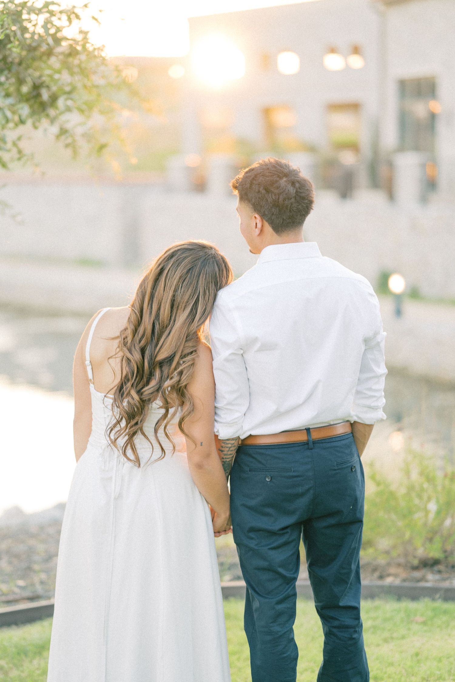A couple stands hand in hand facing a glowing sunset near a peaceful waterway during engagement photos.