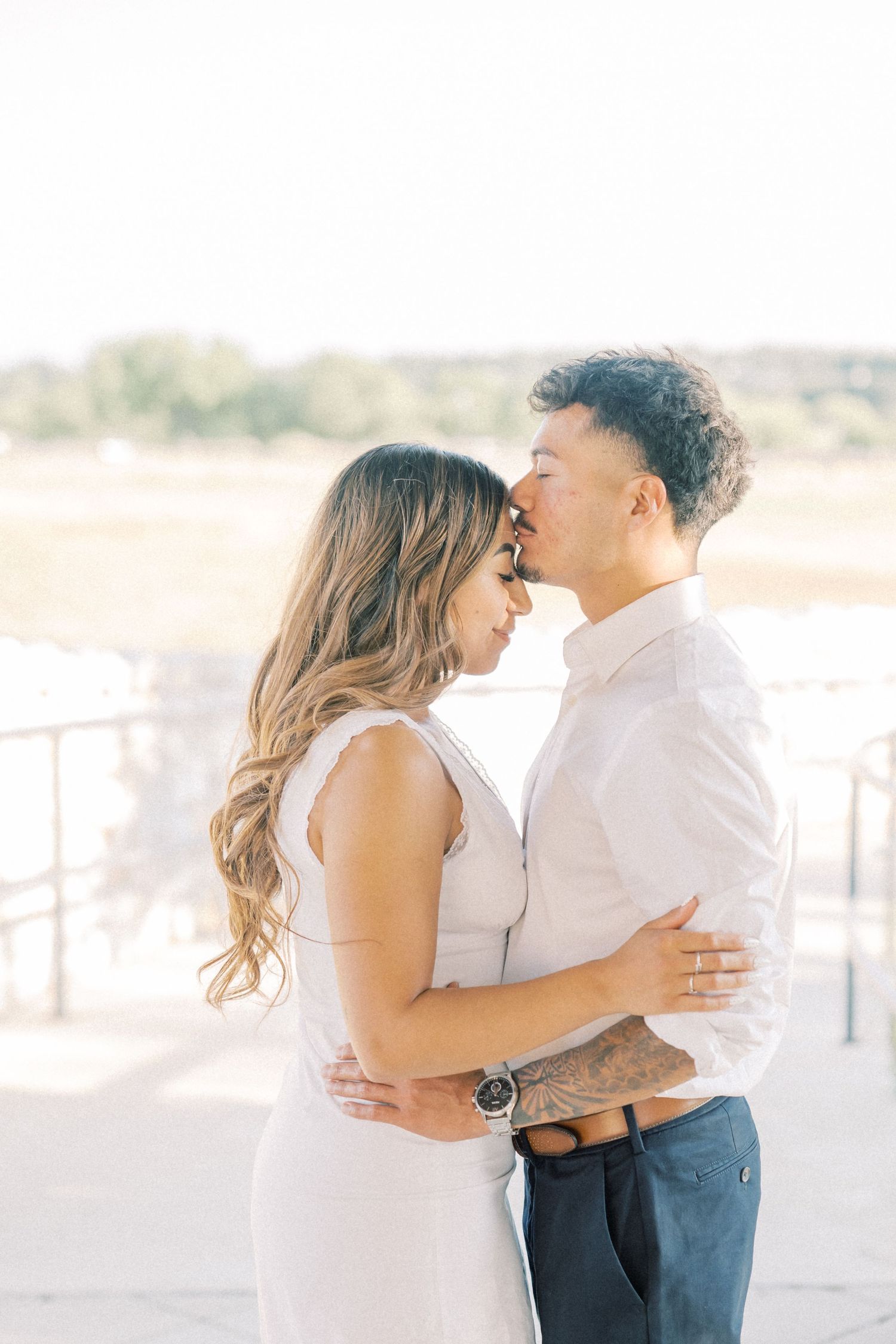 A couple shares a romantic almost-kiss embrace outdoors, dressed in white and navy, with soft natural lighting.