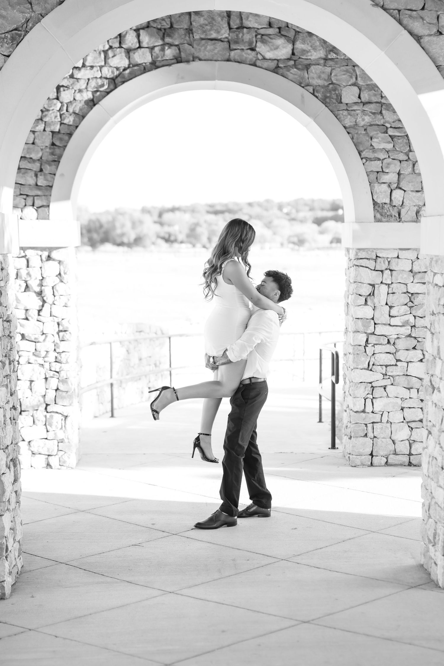 Black and white photo of man lifting woman under stone arch during engagement shoot.
