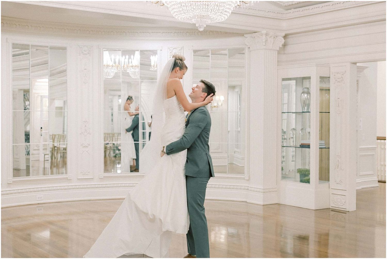 A couple dances in an elegant white room with crystal chandeliers and mirrored walls.