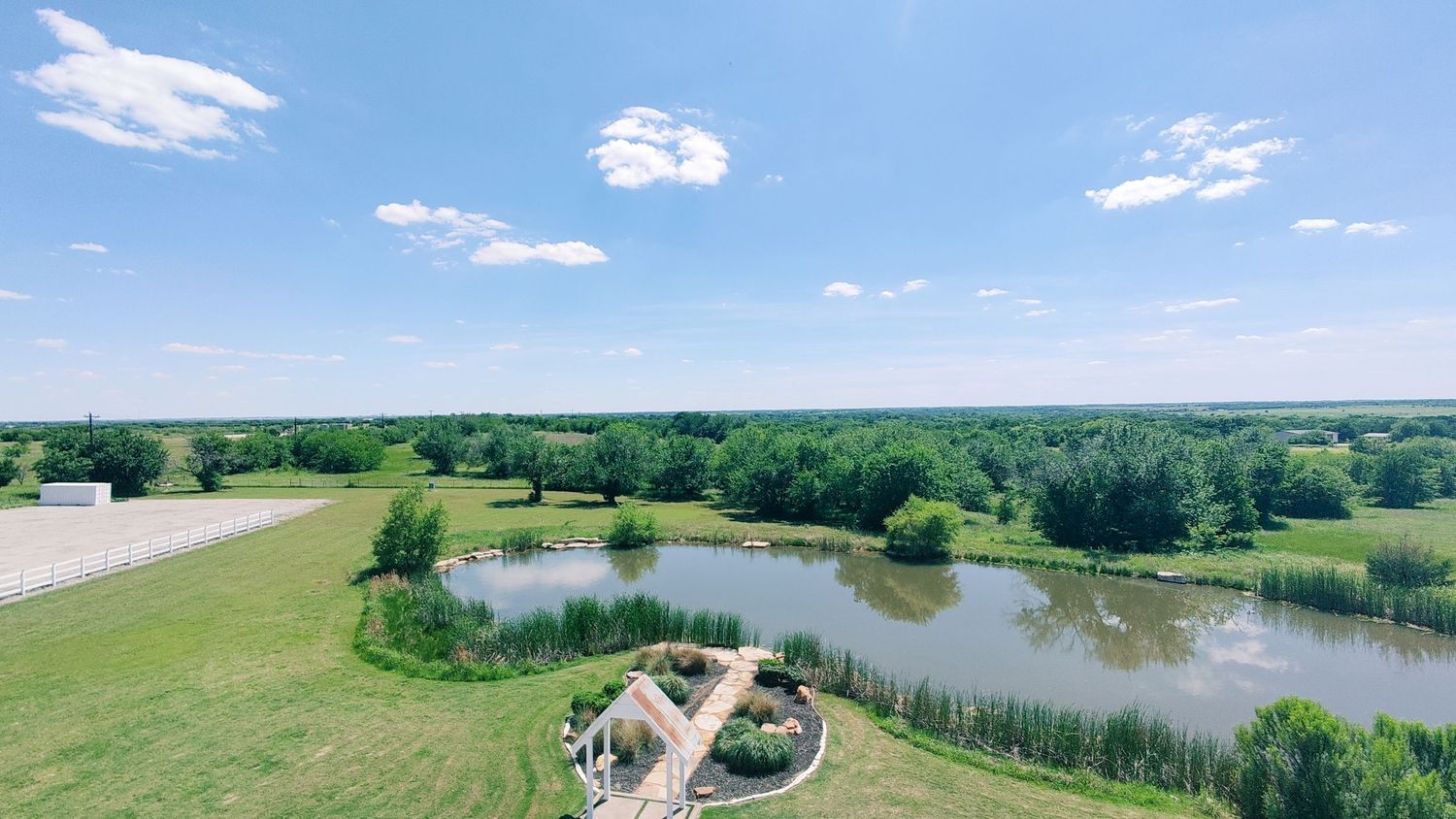Aerial view of a serene rural property featuring a scenic pond, lush green trees, and manicured lawn under a blue sky.