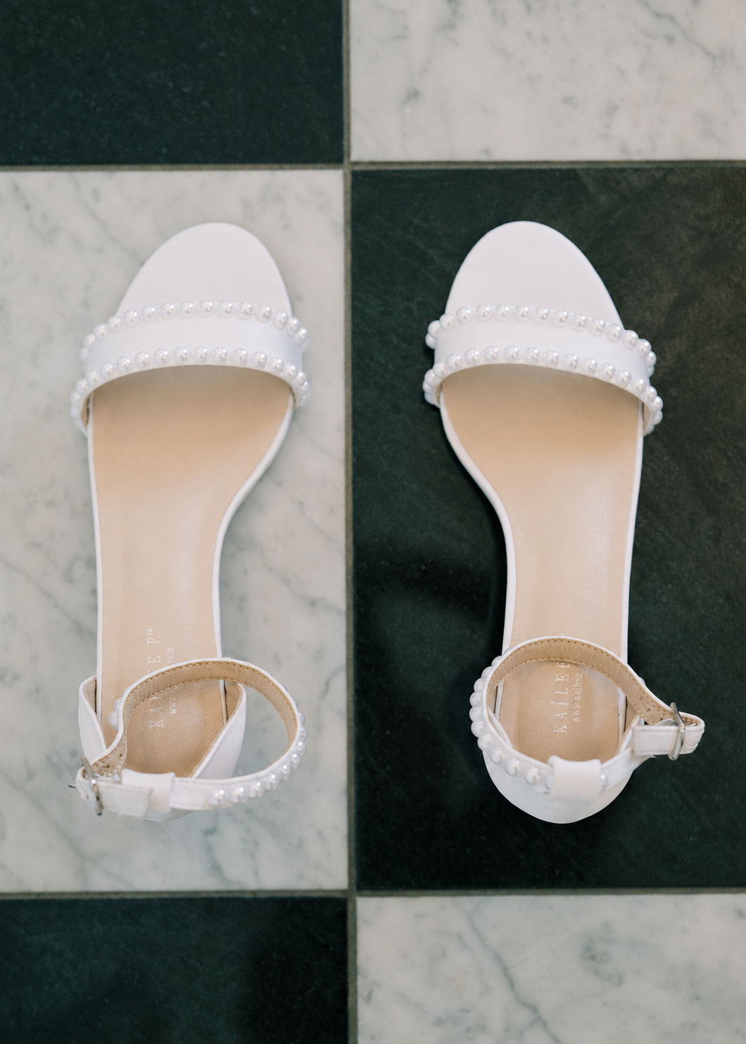 White pearl-embellished bridal heeled sandals with ankle straps displayed on a green and white marble tile floor.